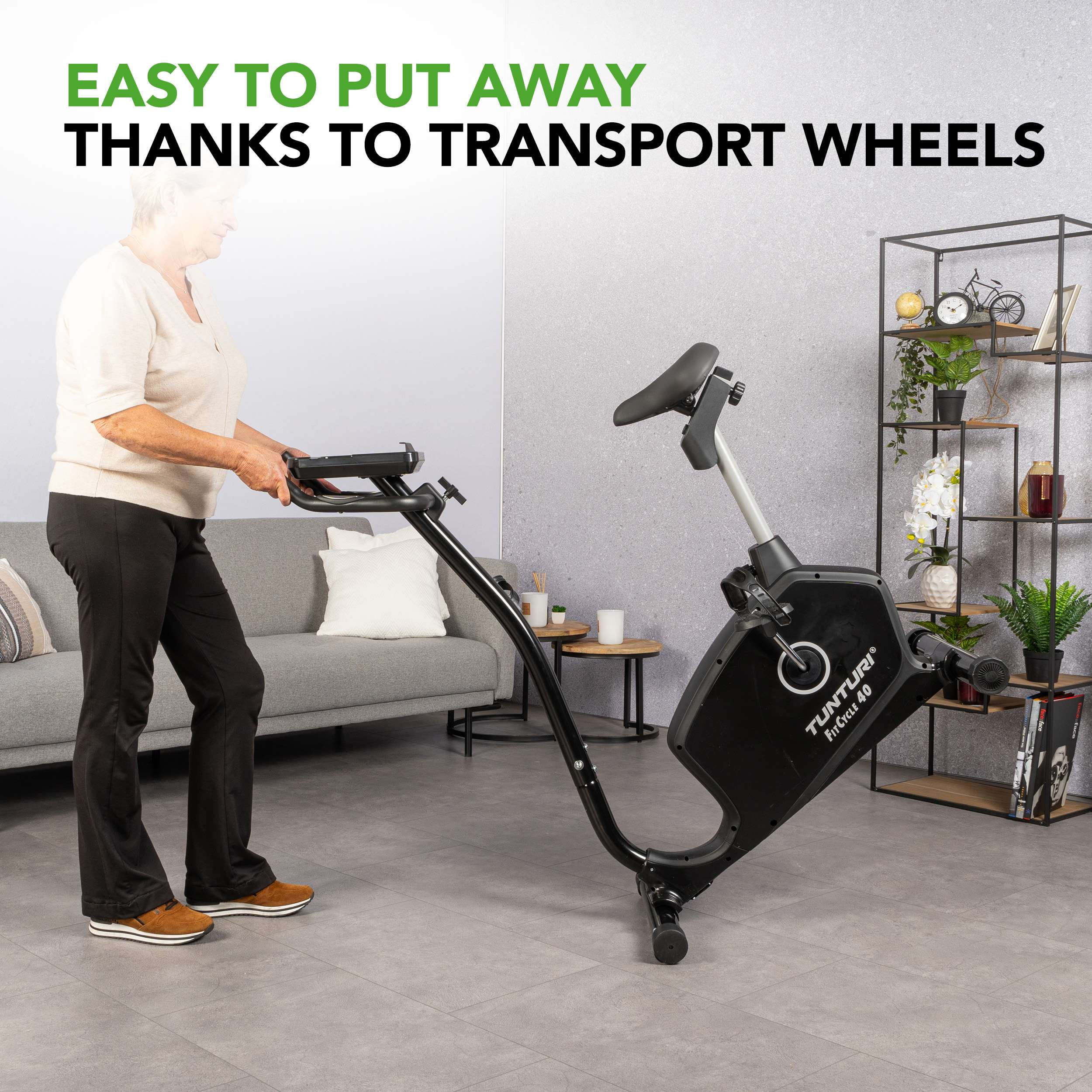 Een oudere vrouw verplaatst een zwarte hometrainer met transportwielen in een woonkamer. De achtergrond omvat een bank en planken.