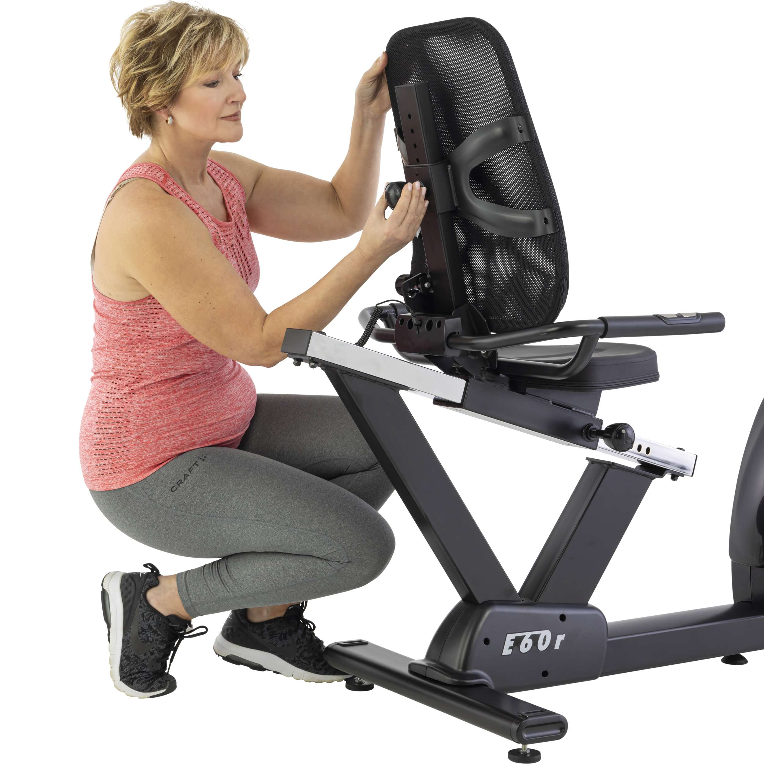 Vrouw die een zwarte stoel van een fitnessfiets verstelt. Ze draagt een roze top, grijze legging en zwarte sneakers. Witte achtergrond.