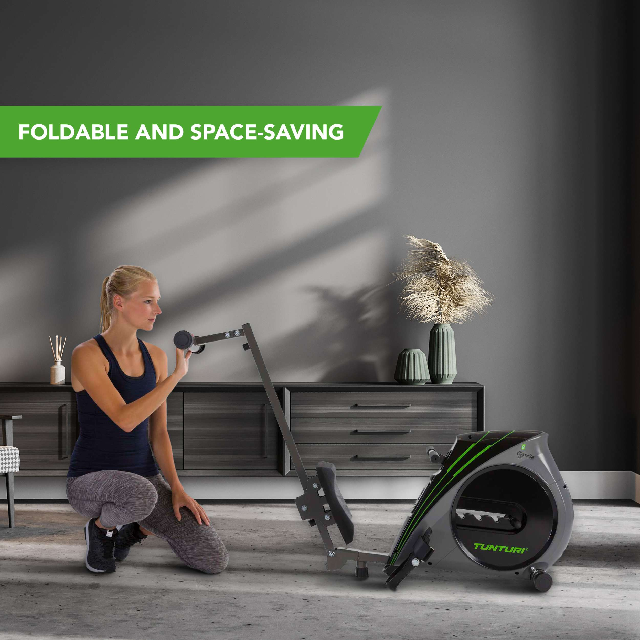 Een vrouw knielt naast een grijszwarte roeimachine. Groene tekst: 'Foldable and Space-saving'. Achtergrond: grijze muur en kast.