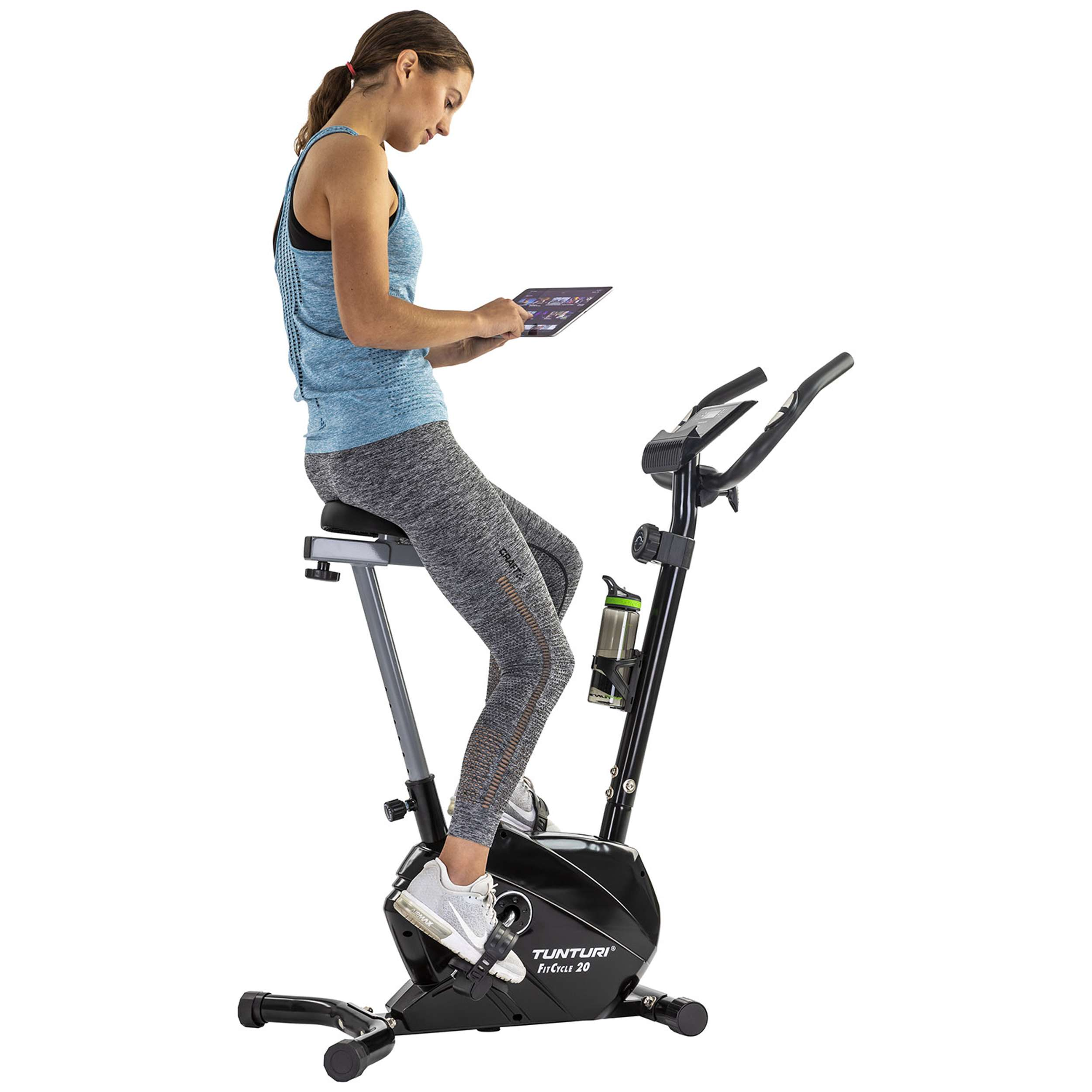Woman on exercise bike, looking at a tablet. She wears a blue top and grey leggings. The bike is black.