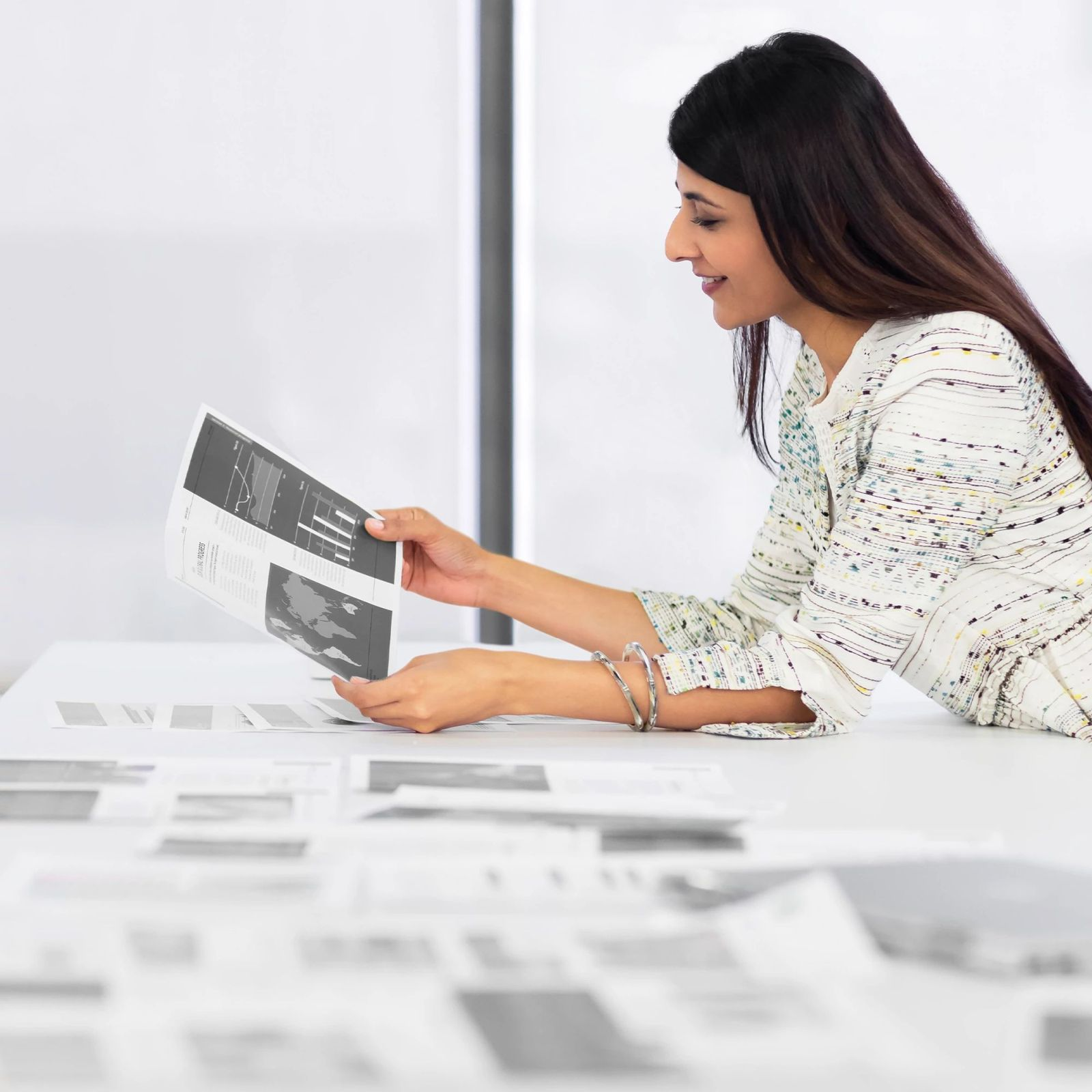 Kobieta przegląda dokument z wykresami. Ma na sobie bluzkę we wzory i bransoletki. Na stole leżą papiery.