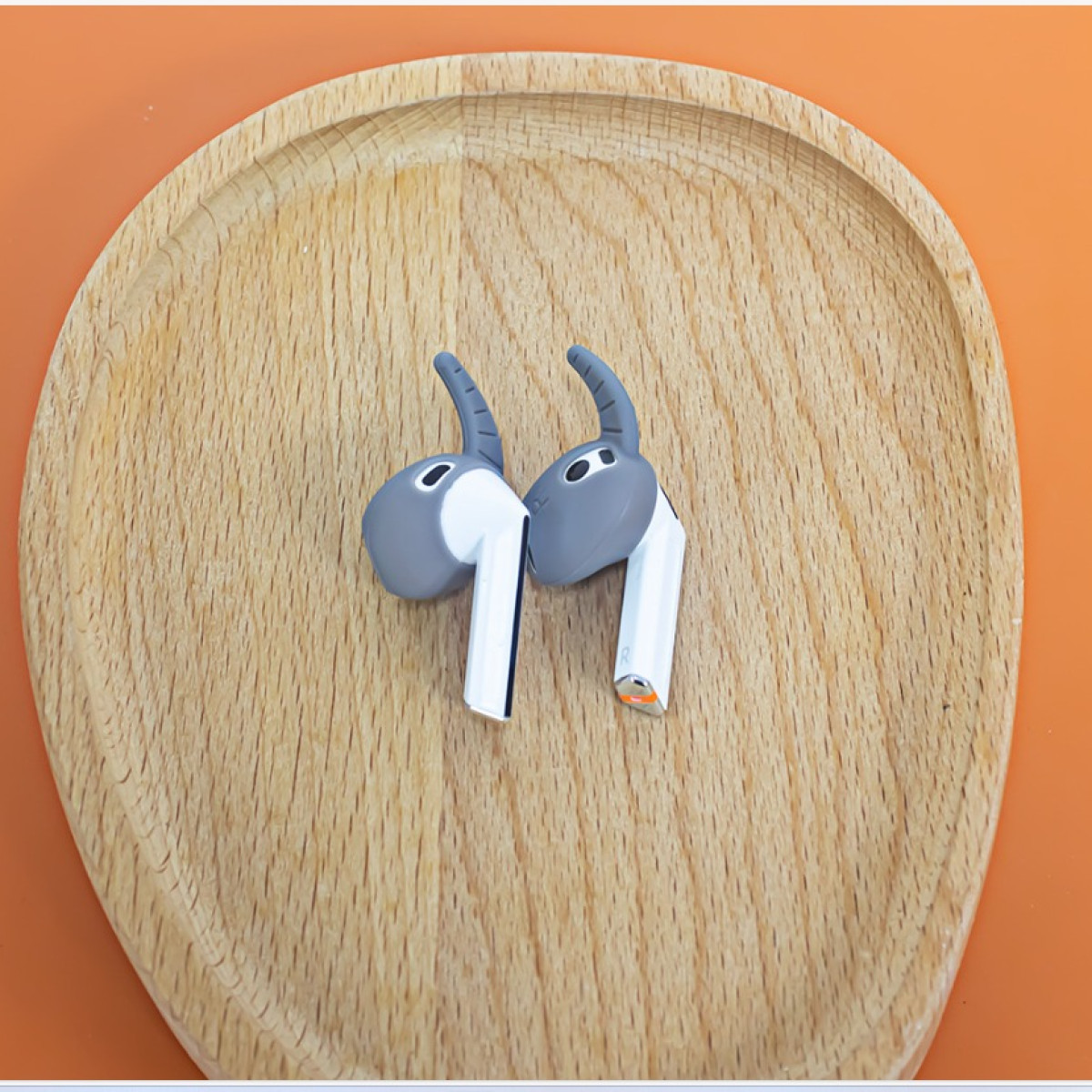 Zwei graue und weiße Ohrhörer mit Silikonhüllen auf einem Holztablett, orangefarbener Hintergrund.