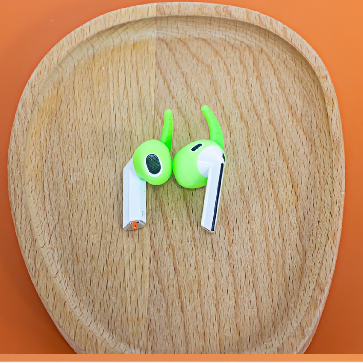 Zwei grüne und weiße Ohrhörer mit Silikonspitzen liegen auf einem Holztablett vor einem orangefarbenen Hintergrund.