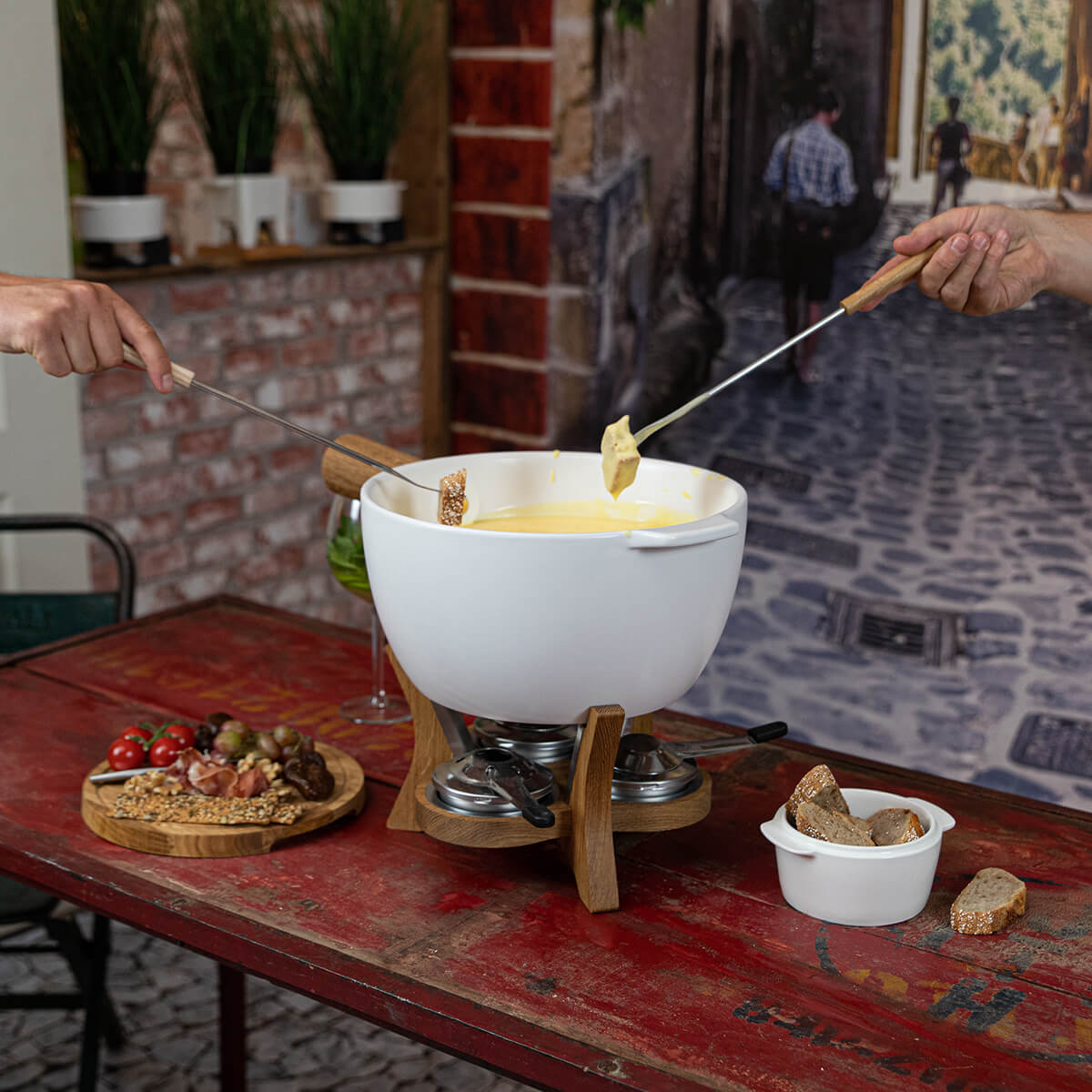 Fondue set on red table. Hands dip bread into cheese. Includes bread, tomatoes, grapes, and meat on wooden plate.