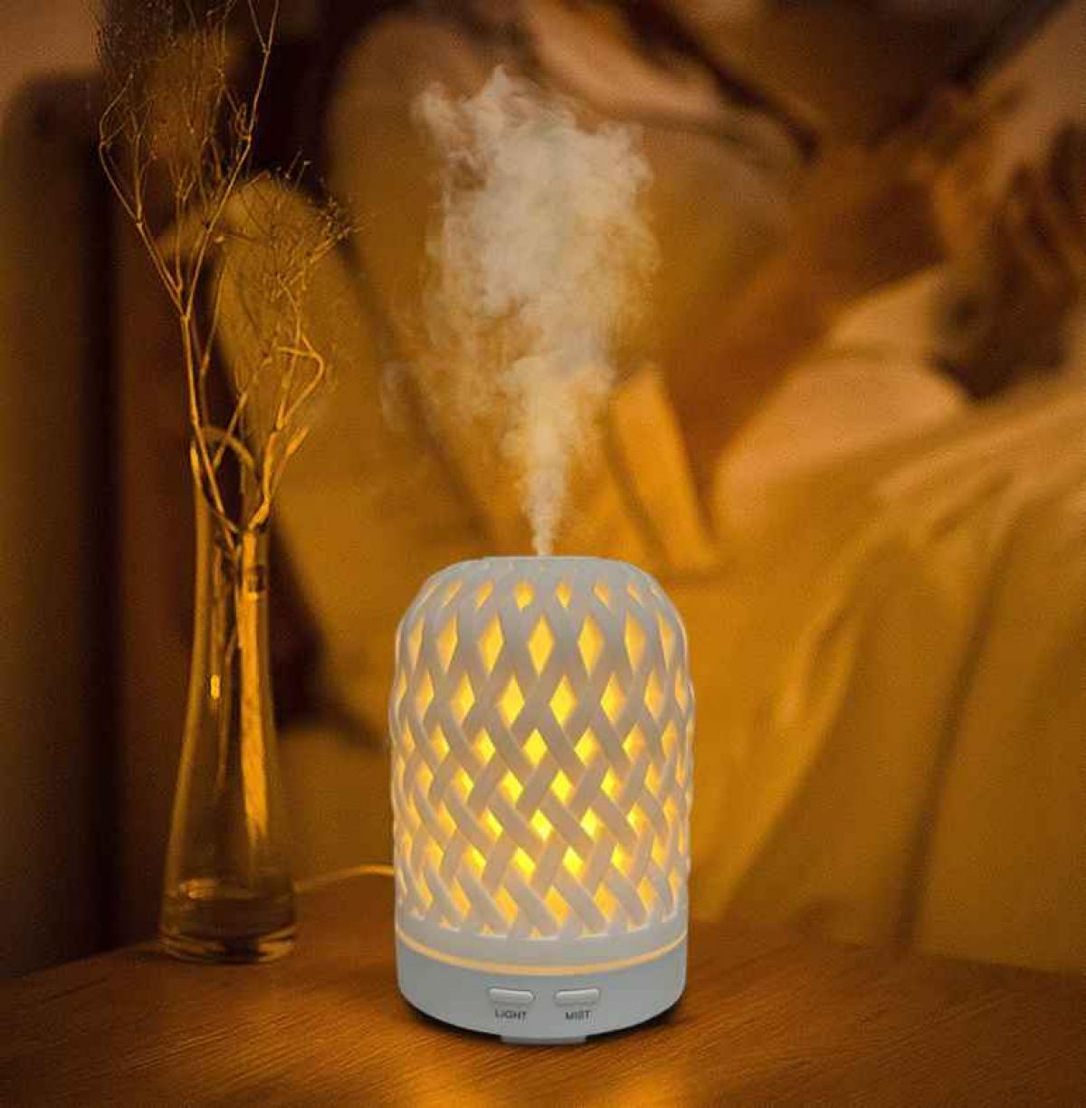 Ein weißer Diffusor mit Gittermuster verströmt Nebel. Er steht auf einem Tisch in der Nähe einer Vase mit getrockneten Ästen. Weiches Licht beleuchtet die Szene.