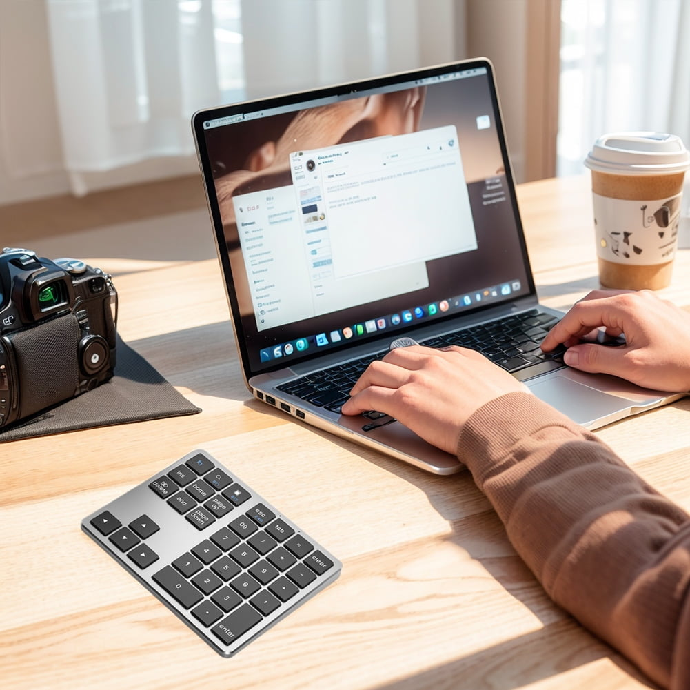 Eine Person benutzt einen Laptop an einem Holztisch. Ein Kaffeebecher und eine Kamera sind ebenfalls vorhanden. Ein numerischer Tastenblock befindet sich im Vordergrund.