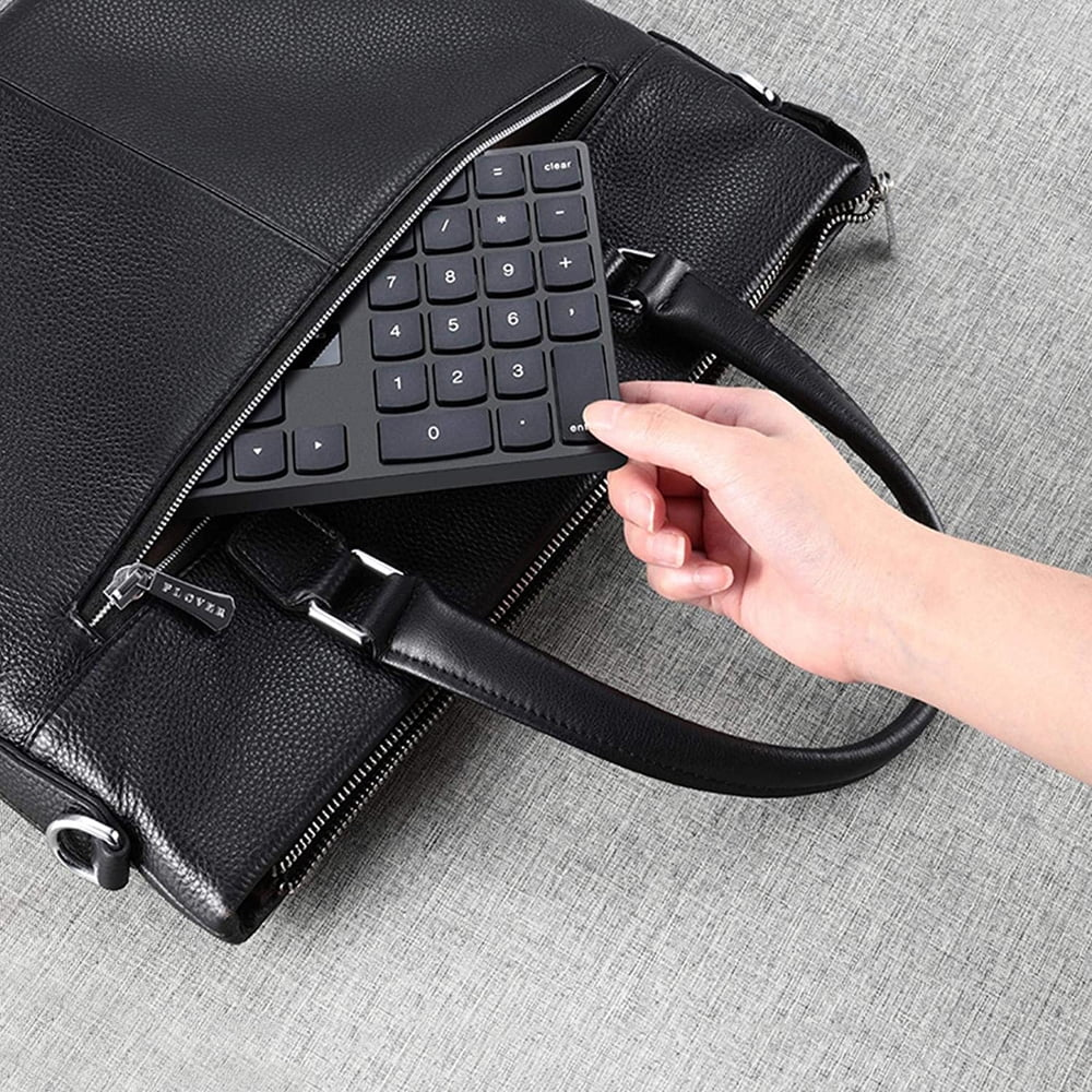 Eine Hand hält einen schwarzen numerischen Ziffernblock, der sich teilweise in einer schwarzen Ledertasche befindet. Die Tasche liegt auf einer grauen strukturierten Oberfläche.