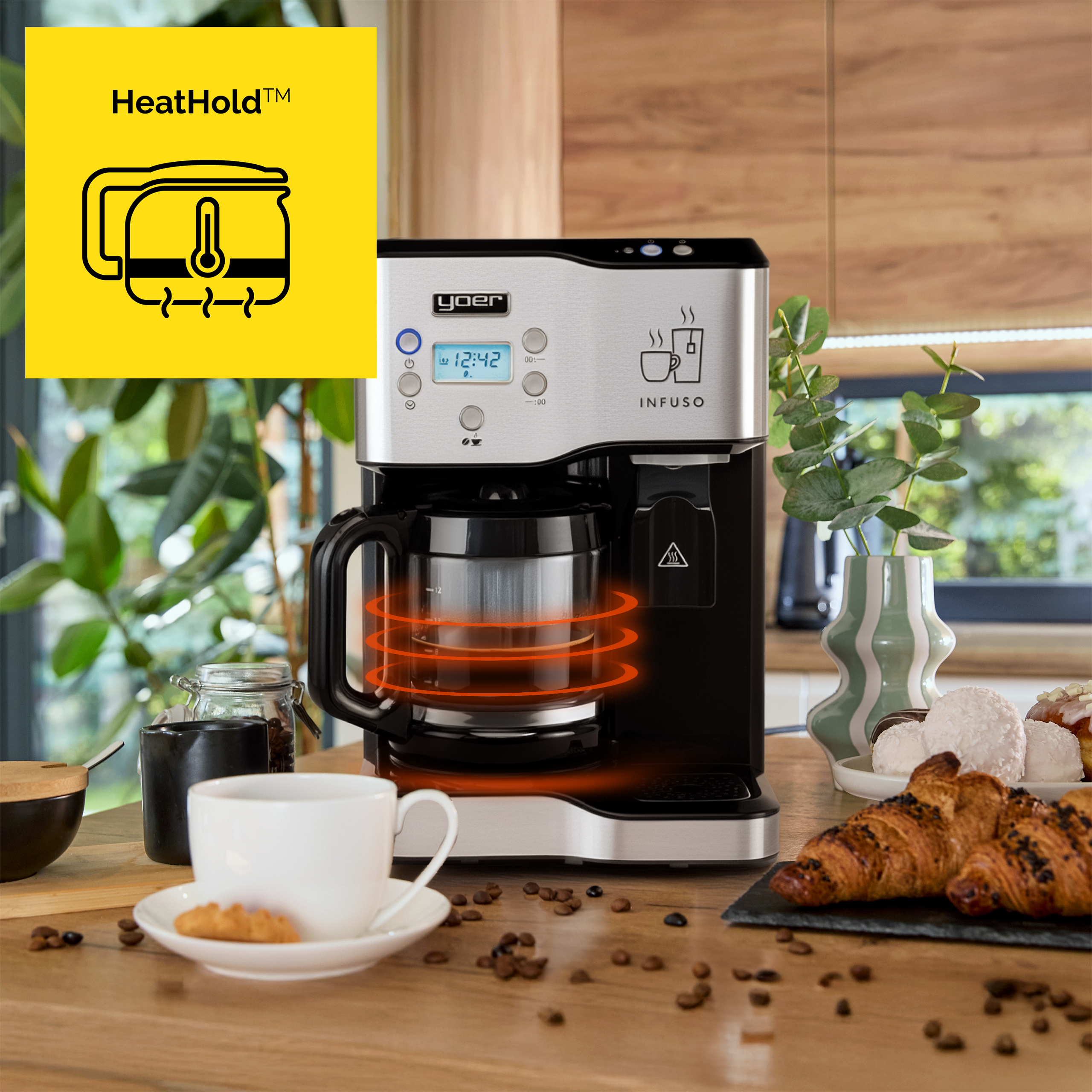 Ekspres do kawy na drewnianym stole z rogalikami, filiżanką i ziarnami. Żółte logo HeatHold po lewej.