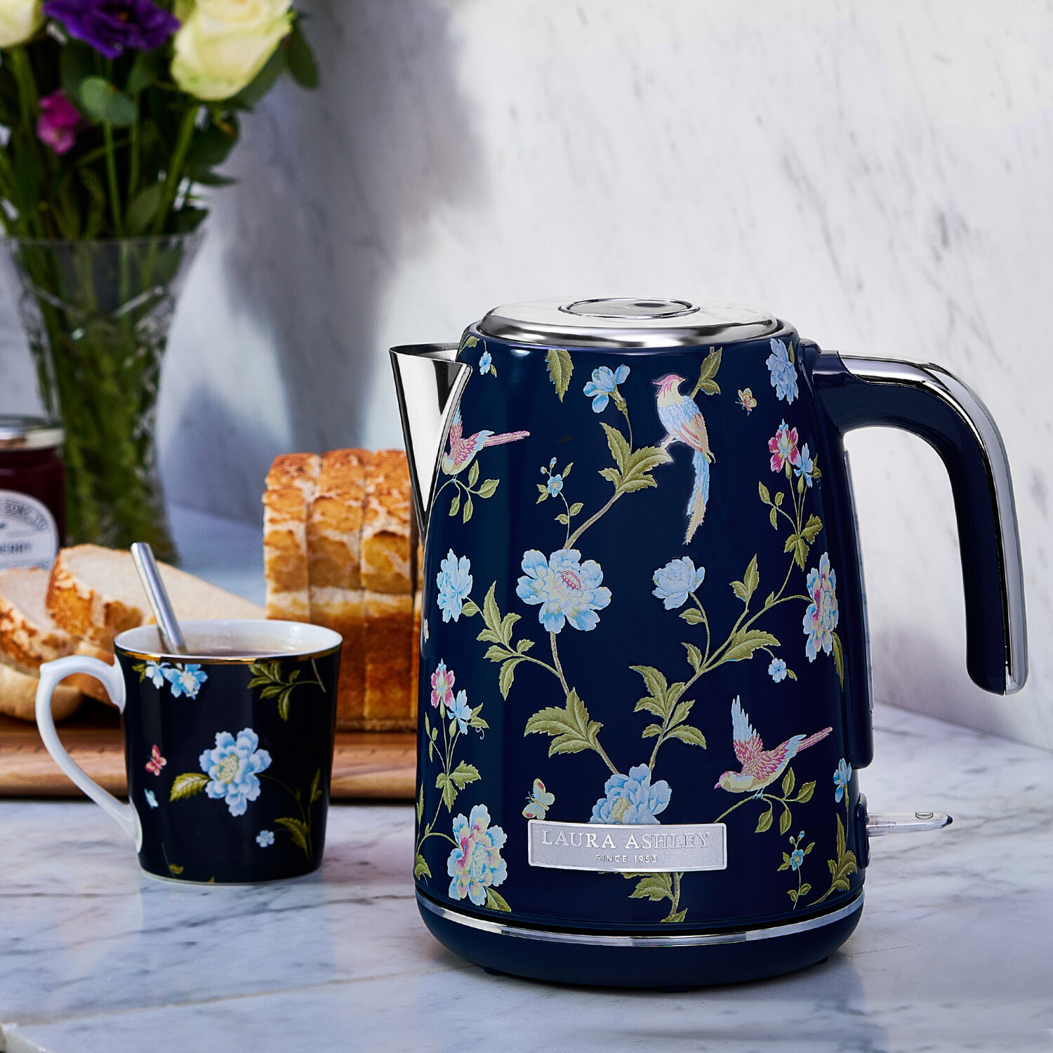 Ein marineblauer Wasserkocher mit Blumen- und Vogelmustern steht auf einer Marmoroberfläche, neben einer passenden Tasse, Brot und Blumen.