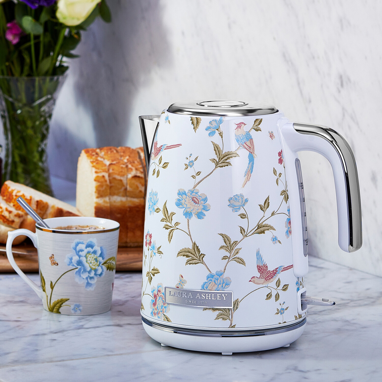 Ein weißer, blumiger Wasserkocher mit passender Teetasse, Brot und Blumen auf einer Marmoroberfläche, Hintergrund ist eine Marmorwand.