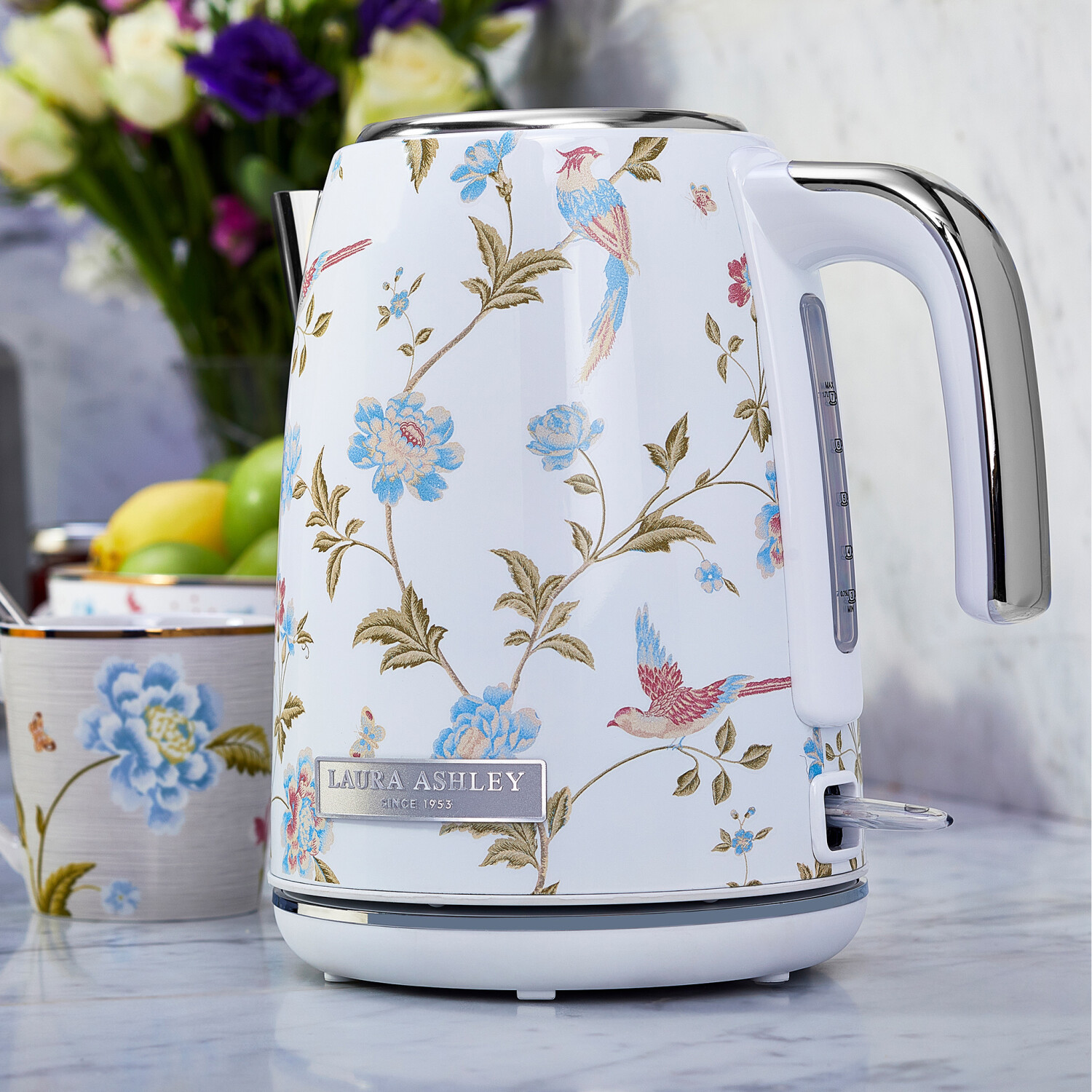 Weißer Wasserkocher mit Blumen- und Vogelmuster, neben einer Tasse und Blumen. Der Wasserkocher hat einen silbernen Griff und ein Laura Ashley-Logo.
