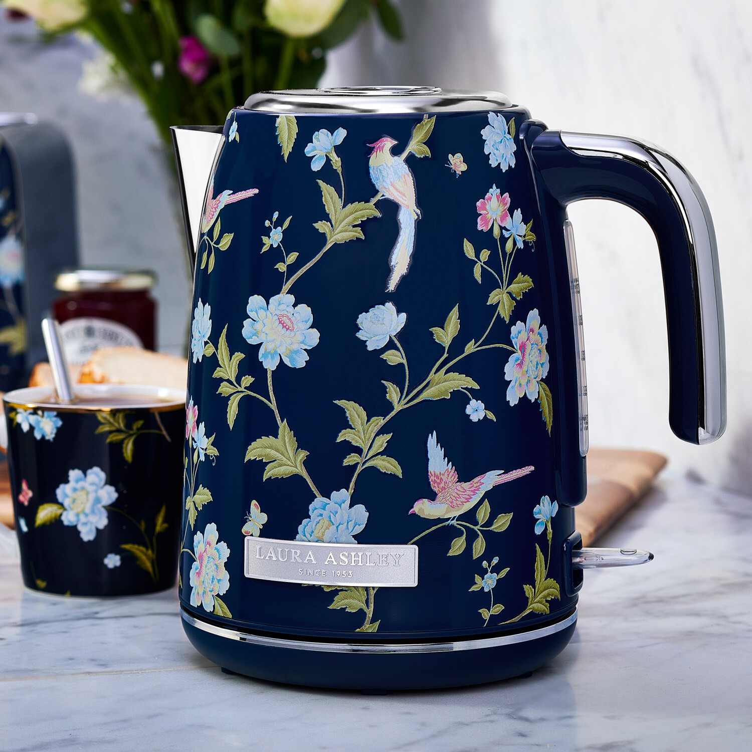 Marineblaues Blumensieb mit Vogel- und Blumendesign. Im Hintergrund sind eine Tasse und Blumen zu sehen. Der Wasserkocher hat einen silbernen Griff.