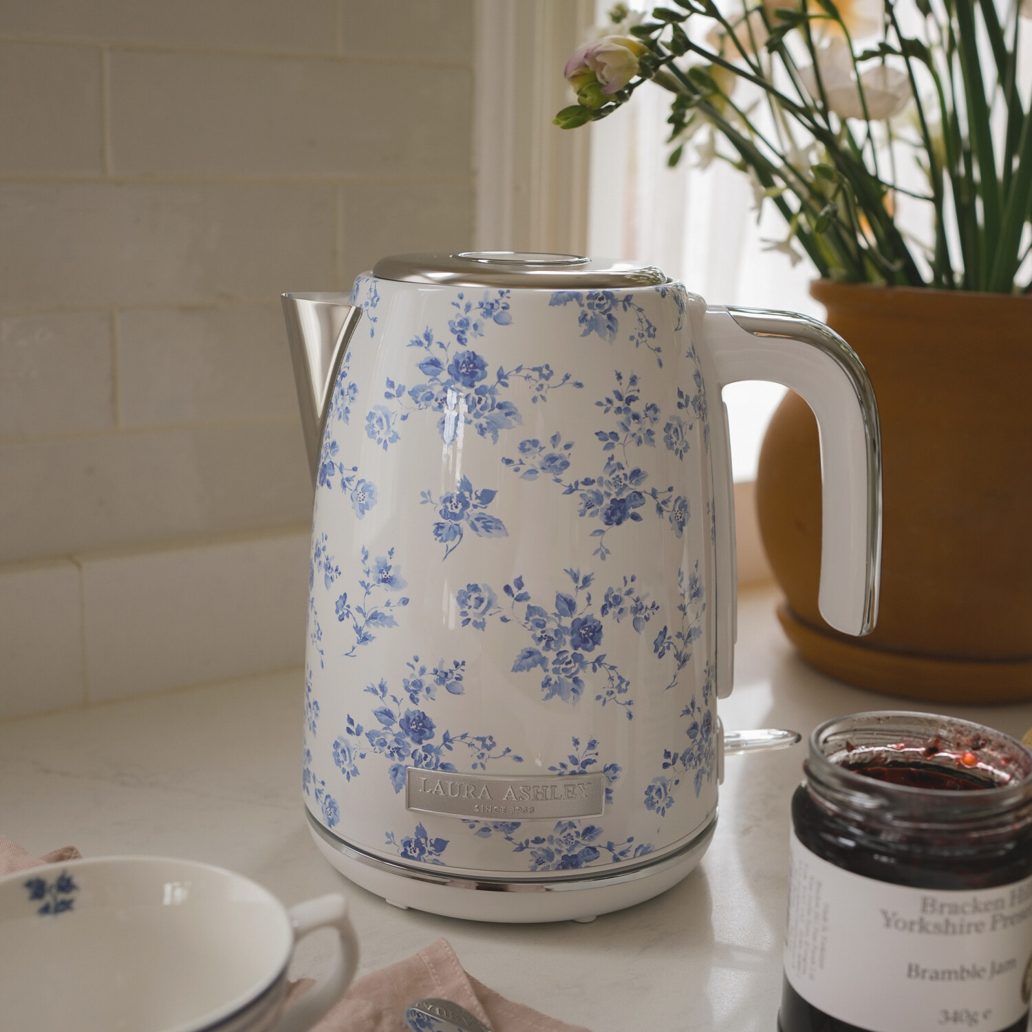 Ein weißer Blumenkessel, eine Tasse und ein Marmeladenglas stehen auf einer Theke. Blumen in einem Topf sind im Hintergrund. Weiches Licht.
