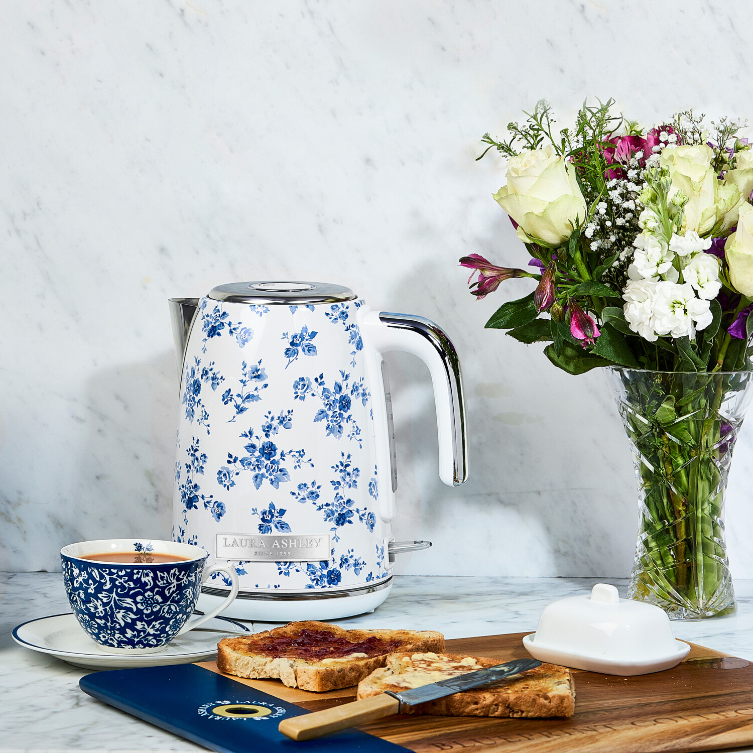 Ein floraler Wasserkocher, eine Teetasse, Toast mit Marmelade und Blumen auf einer Marmoroberfläche. Eine Butterdose und ein Messer sind ebenfalls zu sehen.