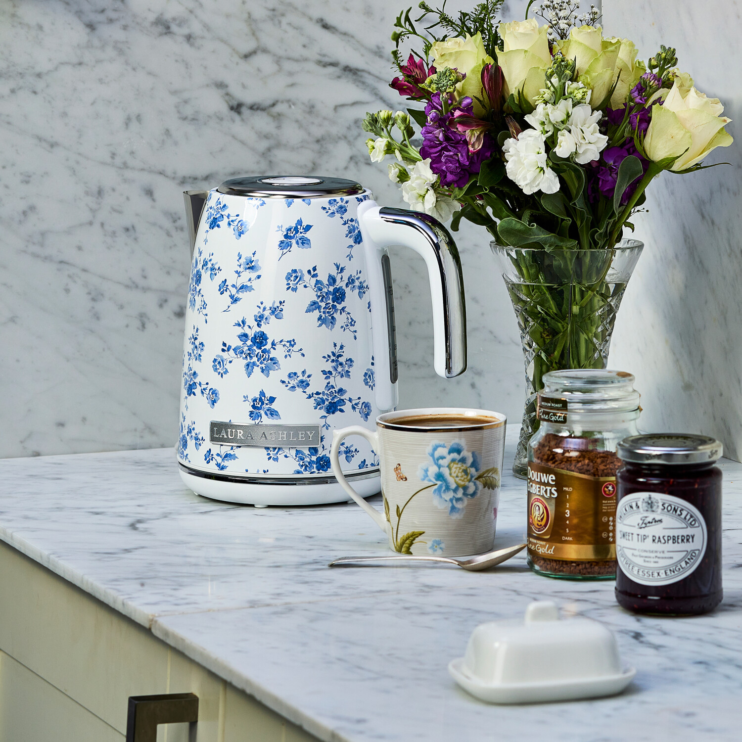 Eine Küchenszene mit einem floralen Wasserkocher, einer Tasse, Blumen, Kaffee, Marmelade und einer Butterdose auf einer Marmorarbeitsplatte.