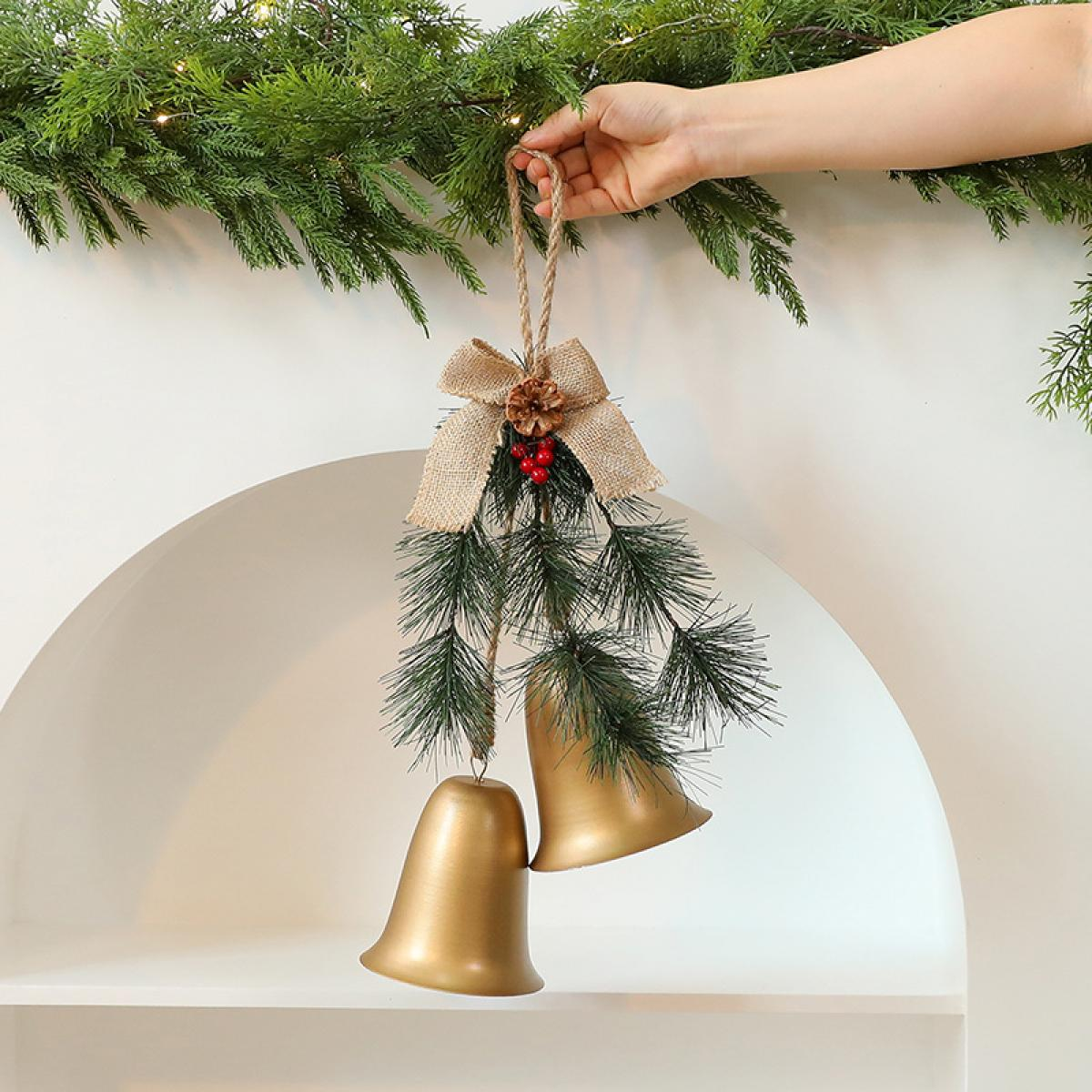 Eine Hand hält eine Weihnachtsdekoration. Sie besteht aus zwei goldenen Glocken, einer Schleife, Tannennadeln und Beeren. Grüner Girlande im Hintergrund.