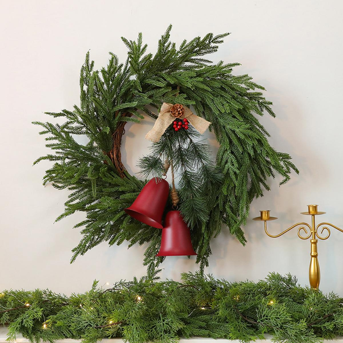 Ein Weihnachtskranz hängt an einer weißen Wand. Er hat rote Glocken, eine Schleife, Tannenzapfen und rote Beeren. Eine Girlande und ein Kerzenständer sind darunter.