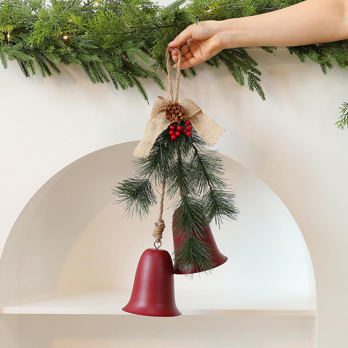 Eine Hand hält eine Weihnachtsdekoration mit zwei roten Glocken, Tannenzweigen, einer Schleife und einem Tannenzapfen.