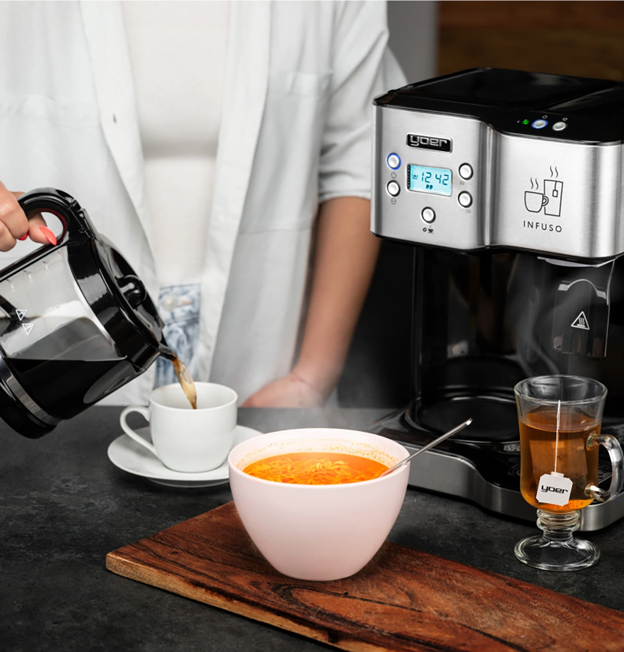 Osoba nalewa kawę do filiżanki. Zupa w misce, herbata w szklance i ekspres do kawy na stole.