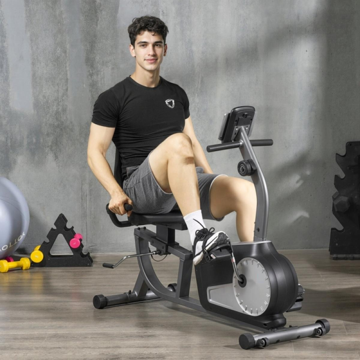 Ein Mann sitzt auf einem Heimtrainer, trägt ein schwarzes Hemd und graue Shorts. Er ist in einem Fitnessstudio.