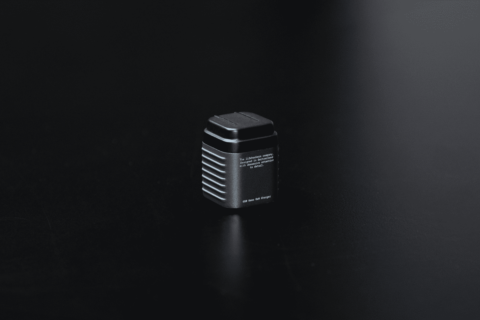 A black 65W GaN charger sits on a dark surface. It has text that reads, 'Designed in Switzerland with obsessive attention to detail.'