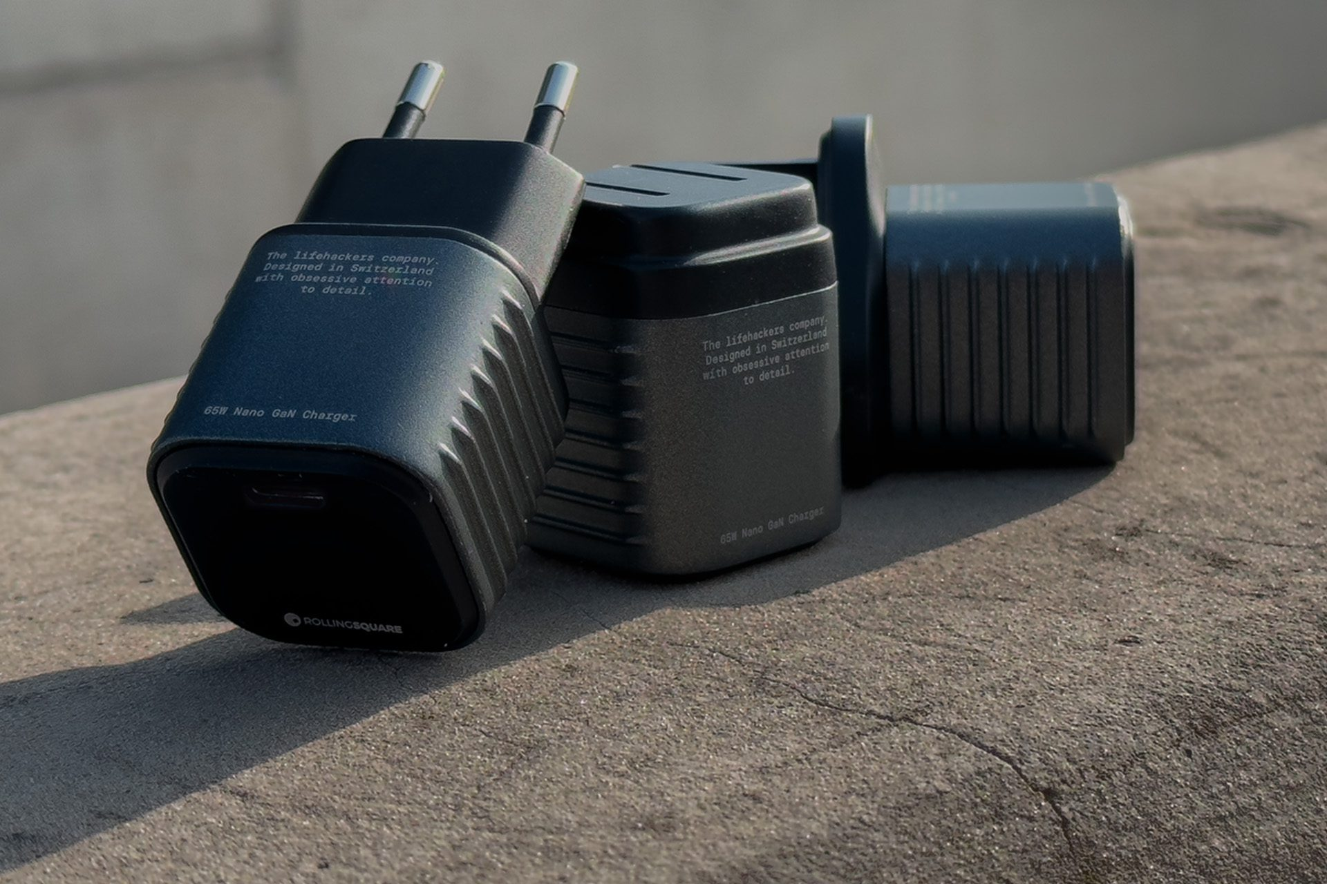 Three dark grey chargers on a stone surface. Each charger has text that reads: 65W Nano GaN Charger and company details.