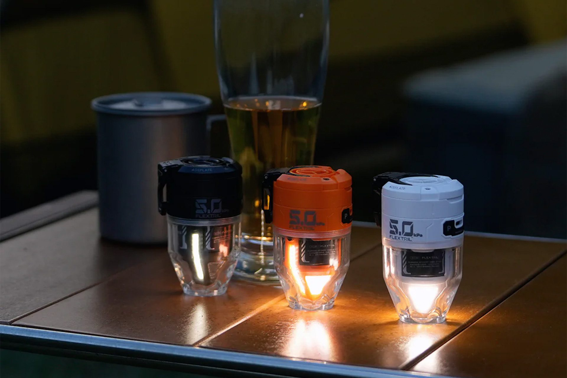 Drei beleuchtete Campinglaternen, schwarz, orange und weiß, auf einem Tisch. Ein Glas Bier und ein Metallbecher sind im Hintergrund.