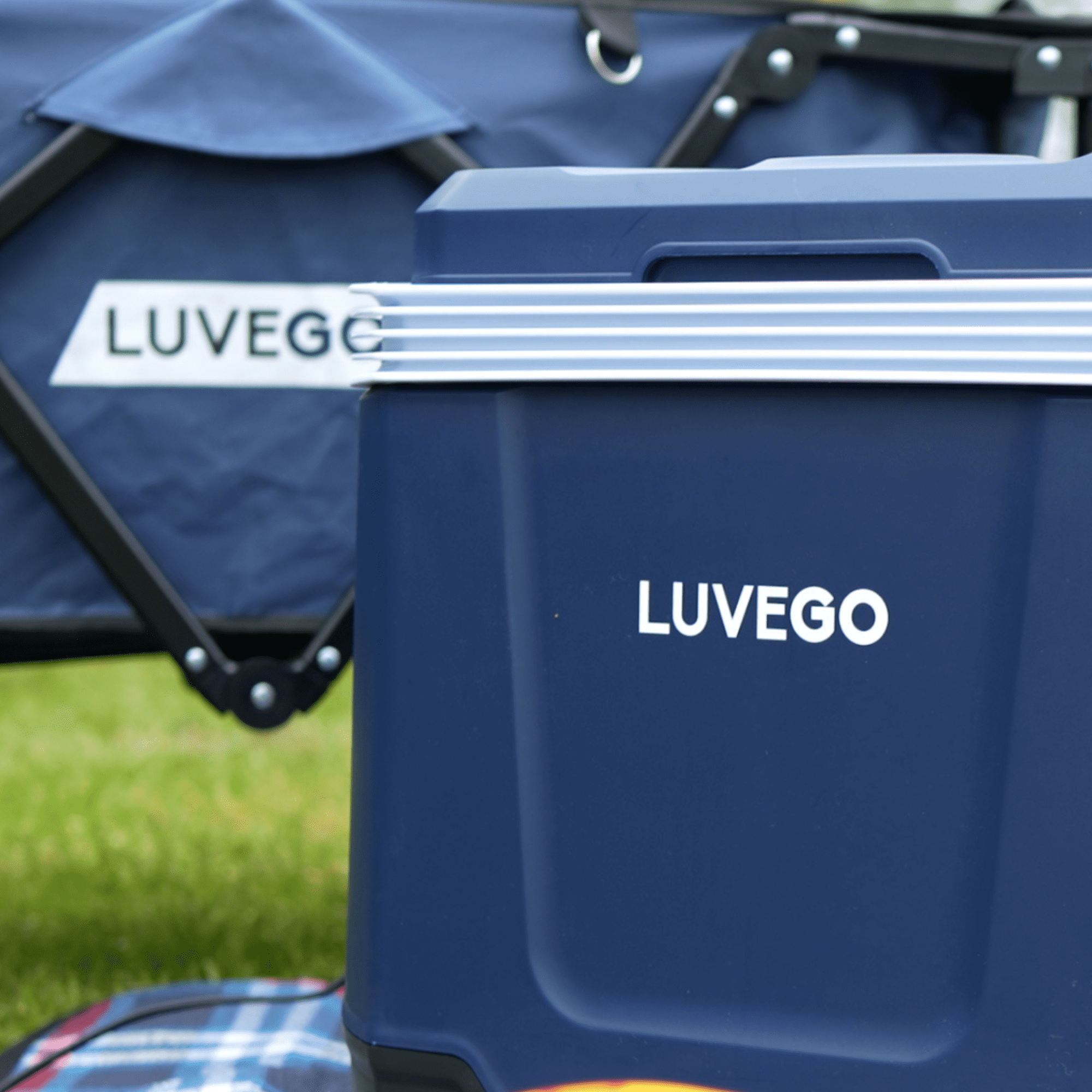 Eine dunkelblaue Kühlbox mit dem Wort Luvgo. Ein blauer Zeltrahmen ist im Hintergrund. Grünes Gras ist sichtbar.