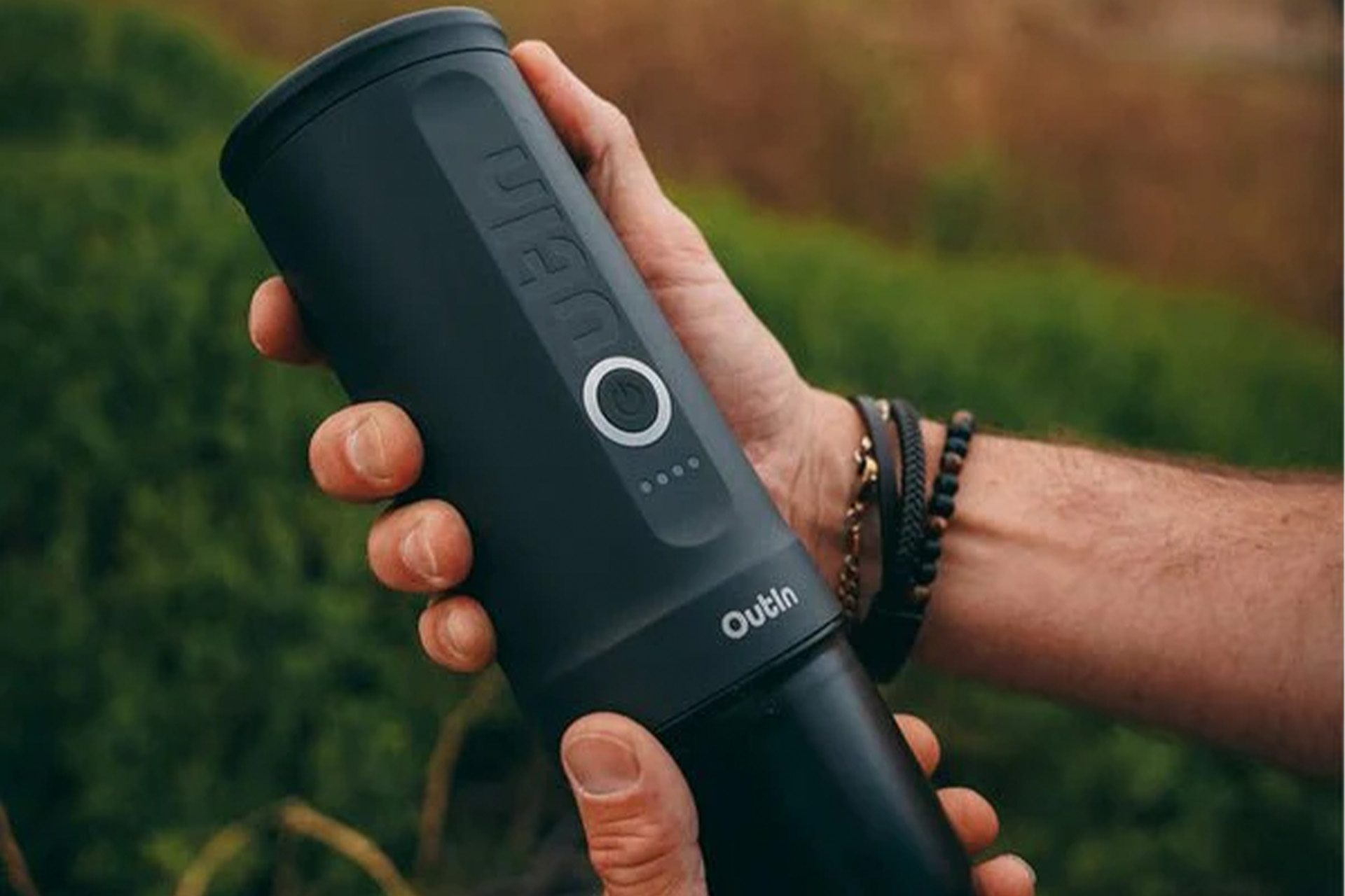 Una mano tiene in mano un dispositivo Outin nero. Ha un pulsante di accensione, spie luminose e il logo Outin. Lo sfondo è verde sfocato.
