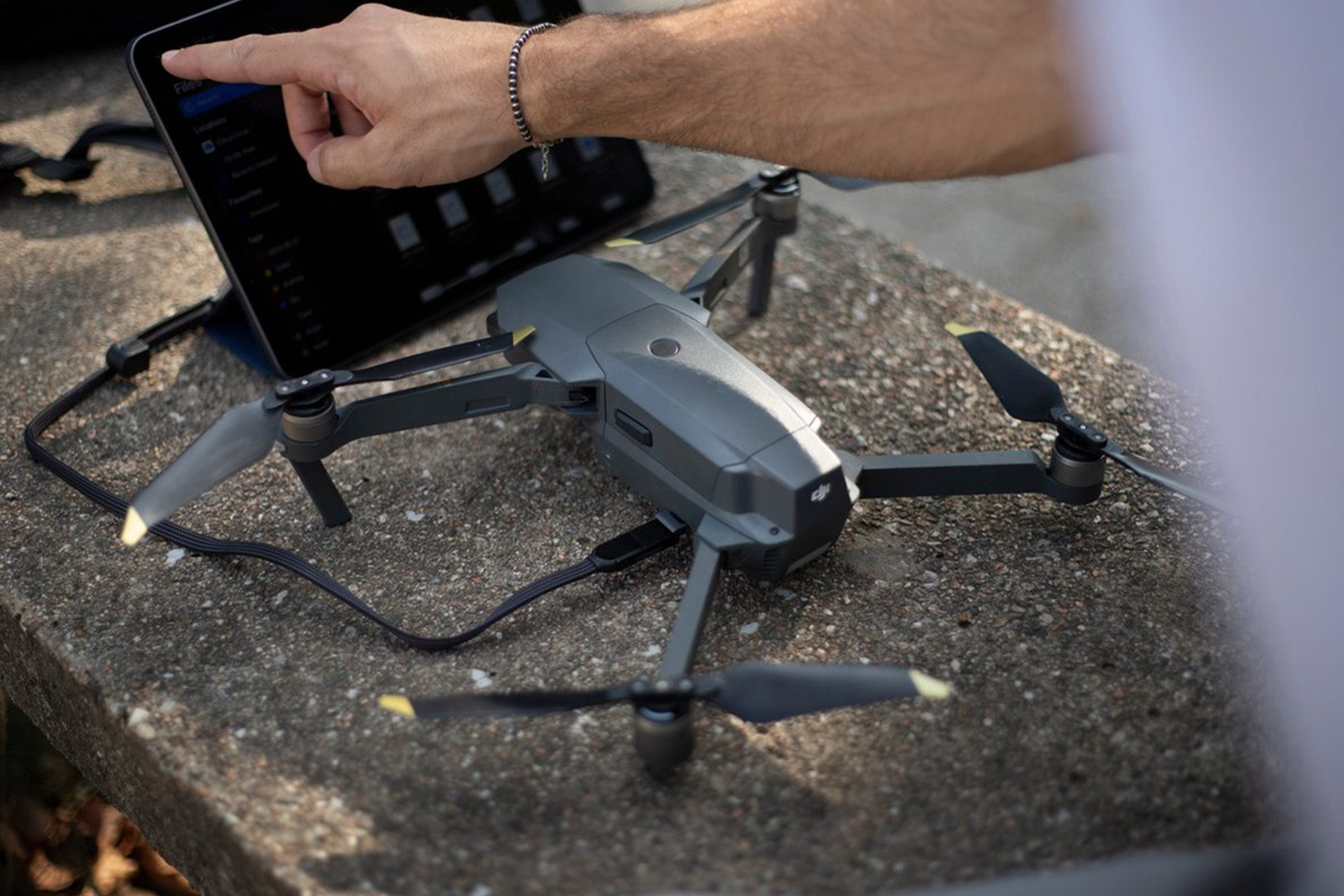 Un drone gris et une tablette sur une surface en béton. Une main pointe vers l'écran de la tablette, un bracelet au poignet.