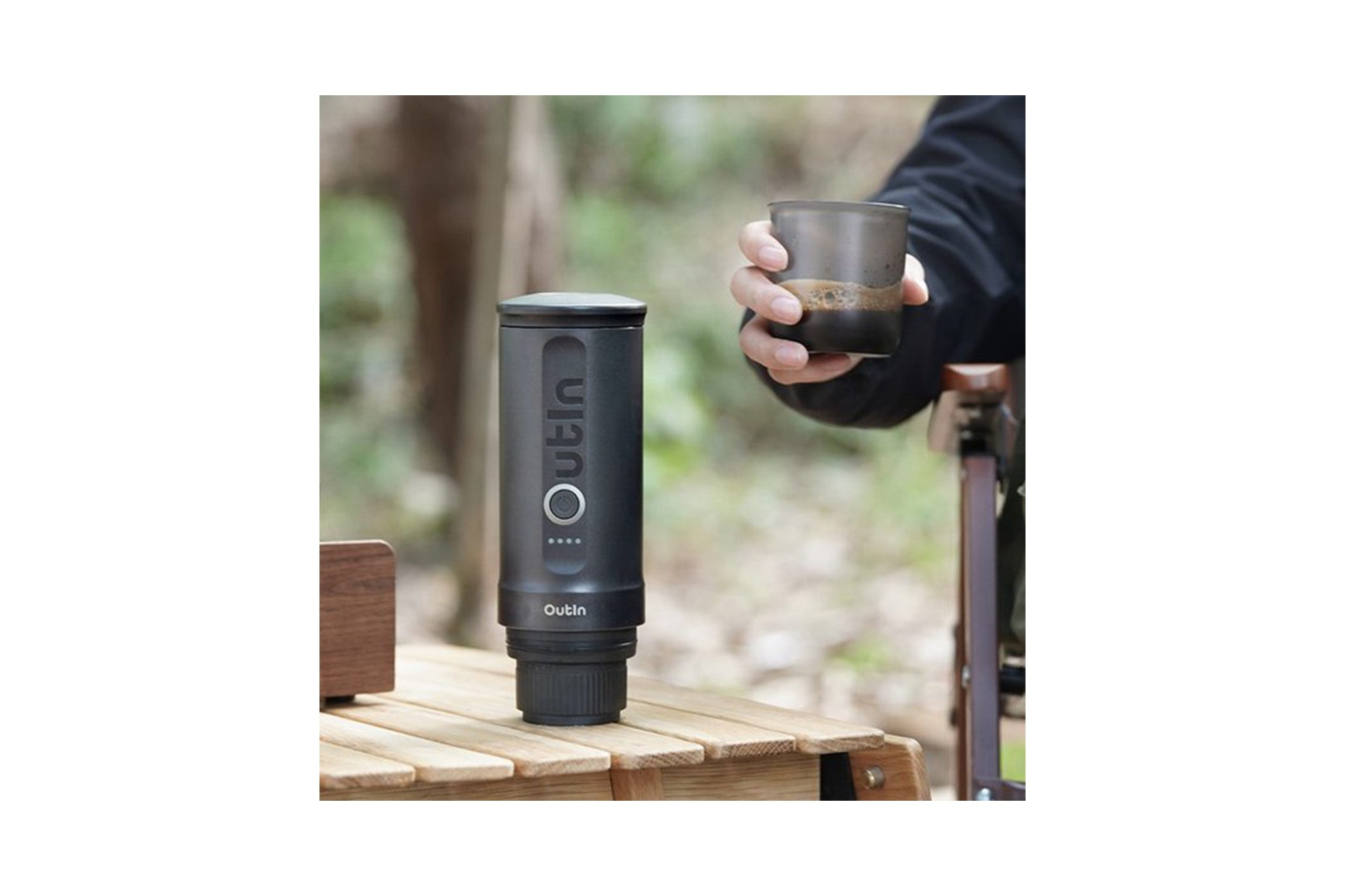 Osoba trzymająca szklankę kawy obok czarnego przenośnego ekspresu do kawy Outin na drewnianym stole.