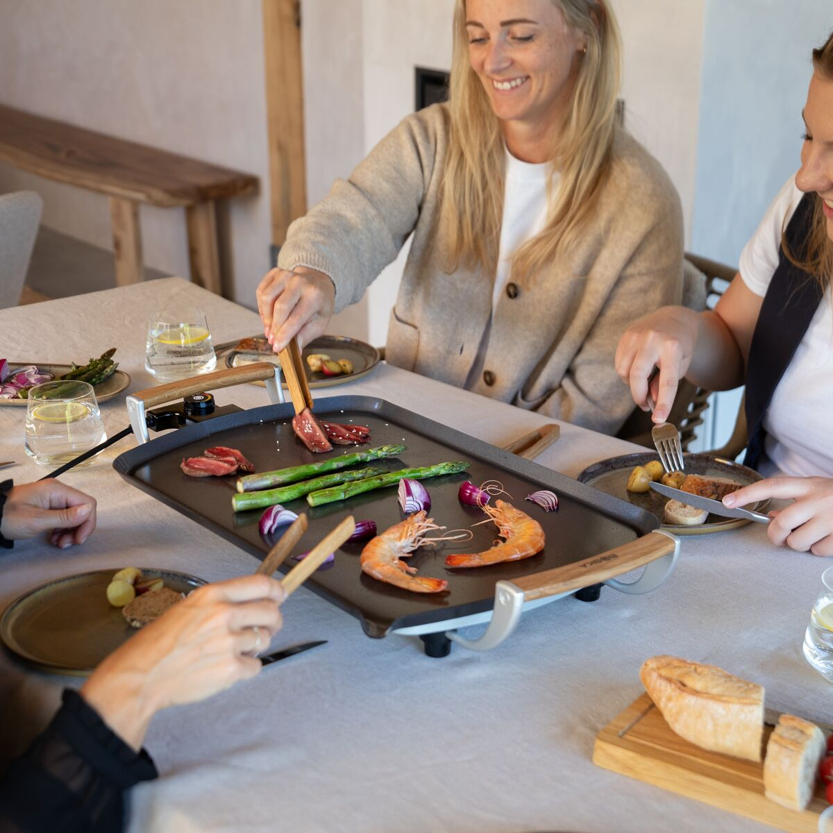People grilling food at a table. Includes meat, asparagus, shrimp, and onion. Smiling women. Plates and glasses are present.