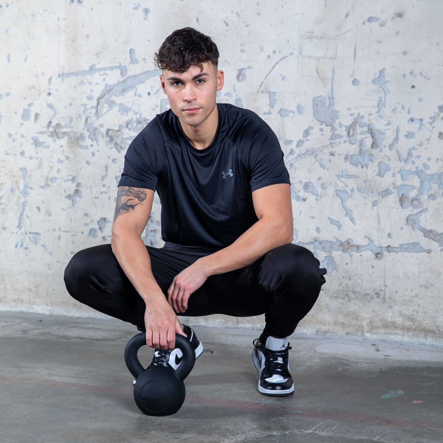 Ein Mann in schwarzer Sportkleidung hockt und hält eine schwarze Kettlebell. Er schaut in die Kamera. Im Hintergrund ist eine Wand.