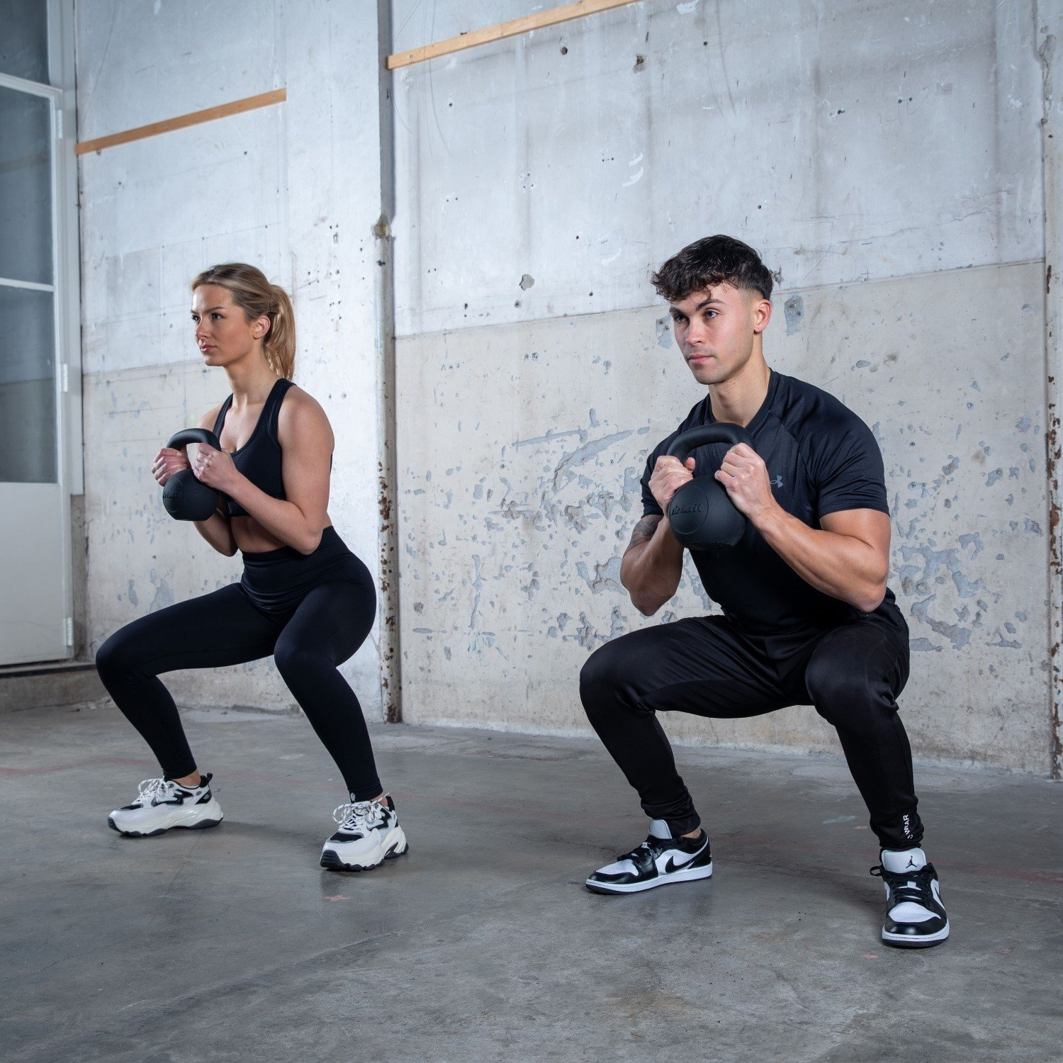 Zwei Personen machen Kniebeugen mit Kettlebells in einem Fitnessstudio. Sie tragen schwarze Trainingskleidung. Der Hintergrund ist eine Betonmauer.