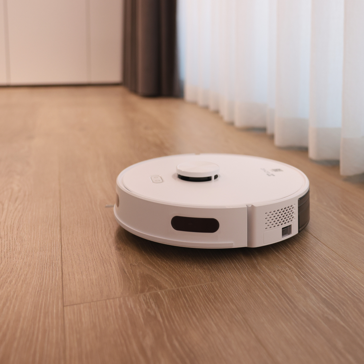 Biały robot odkurzający na drewnianej podłodze. W tle zasłony i ściana.