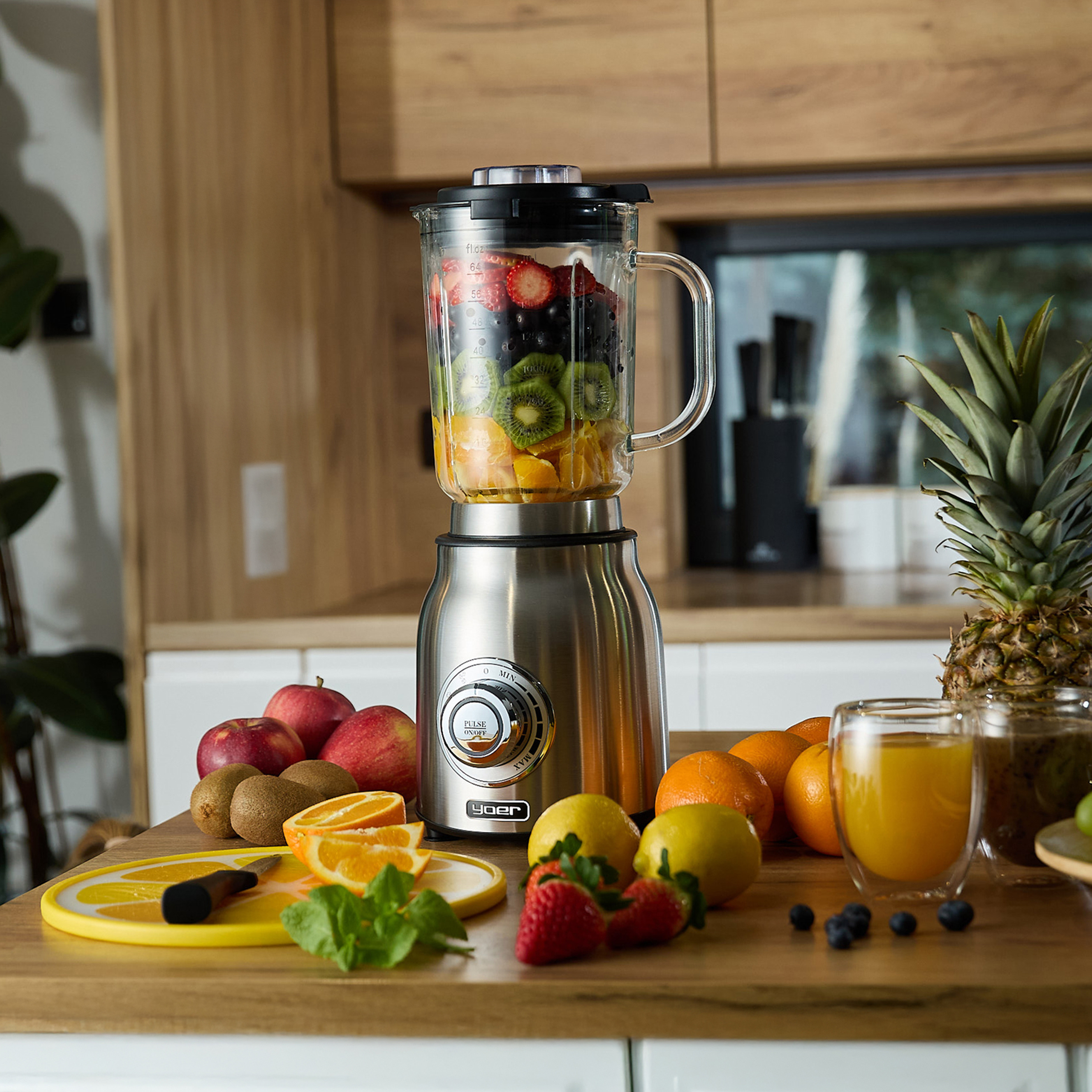 Blender wypełniony owocami, otoczony owocami, sokiem i ananasem na drewnianej powierzchni w kuchni.