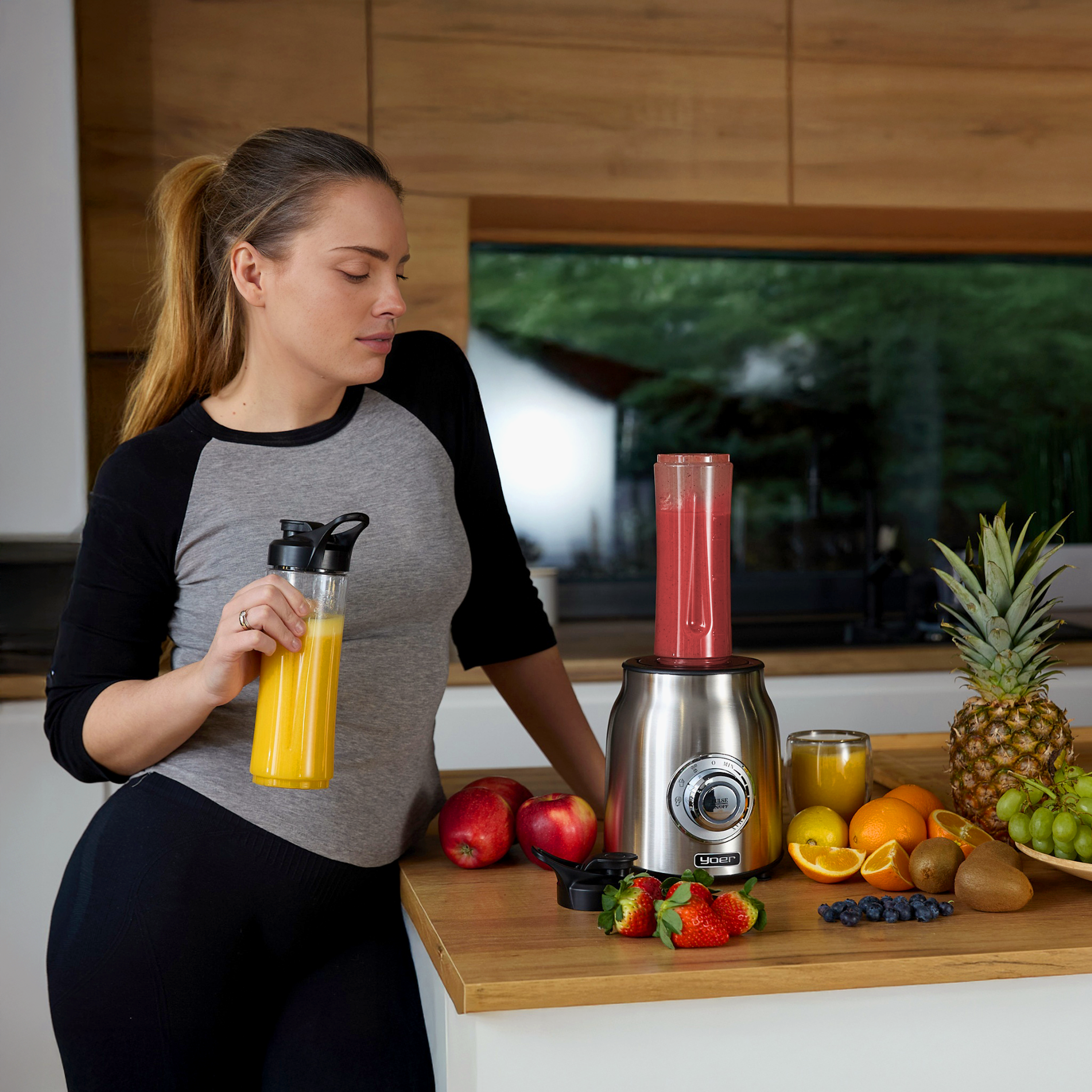 Kobieta z blenderem i owocami. Trzyma butelkę soku. Owoce takie jak jabłka, truskawki i pomarańcze są na stole.