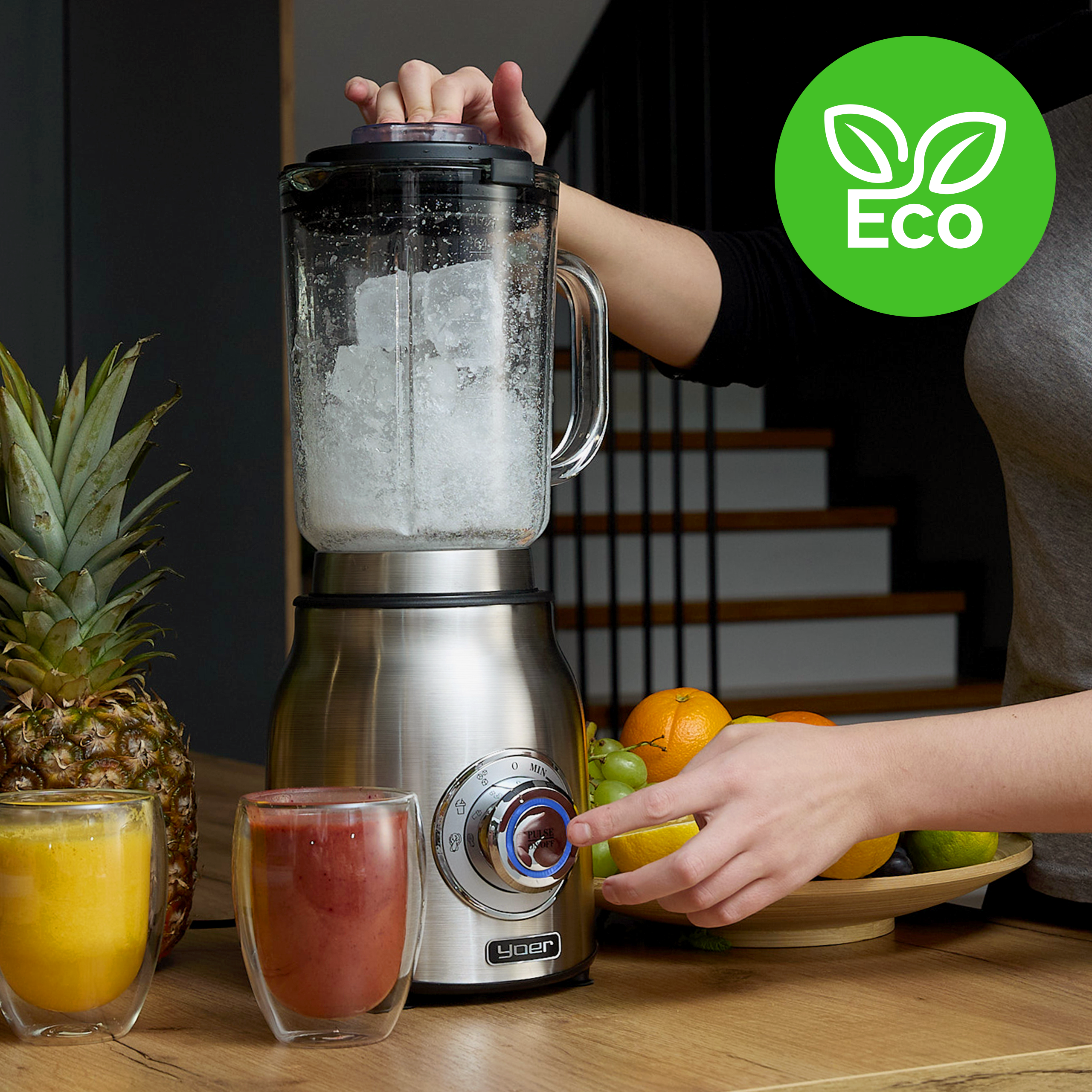 Blender z lodem, szklankami soku, owocami i ananasem na drewnianej powierzchni. Widoczne logo Eco.