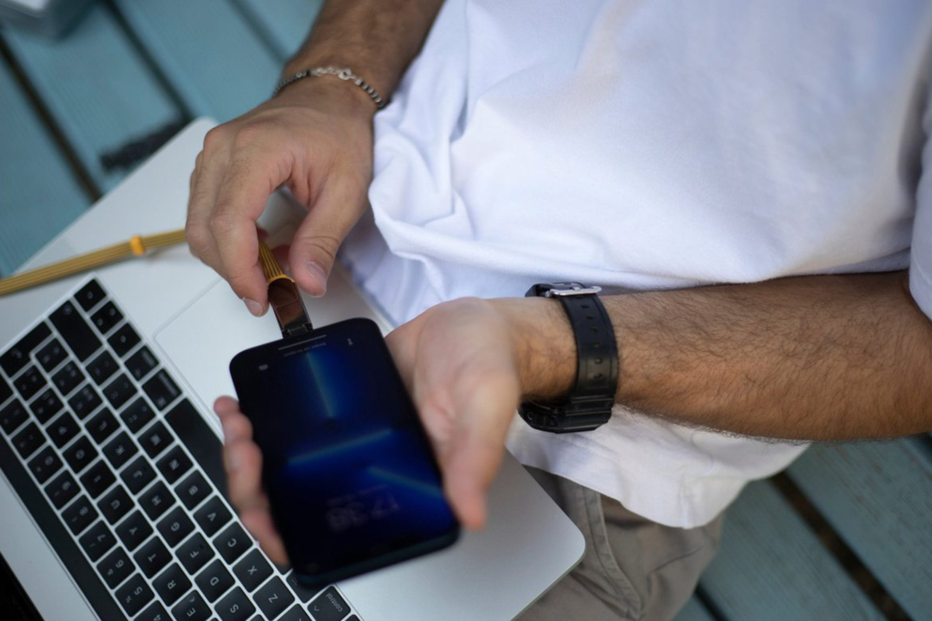 Une personne branche un câble USB sur un smartphone. Un ordinateur portable est à côté du téléphone. La personne porte une montre et un bracelet.