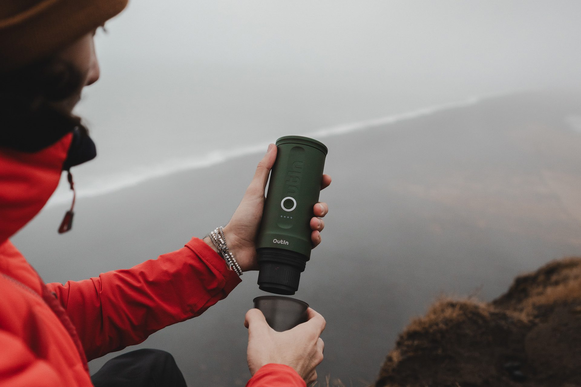 Osoba trzymająca zielony filtr wody nad kubkiem. Nosi czerwoną kurtkę. Zachmurzone niebo i krajobraz w tle.