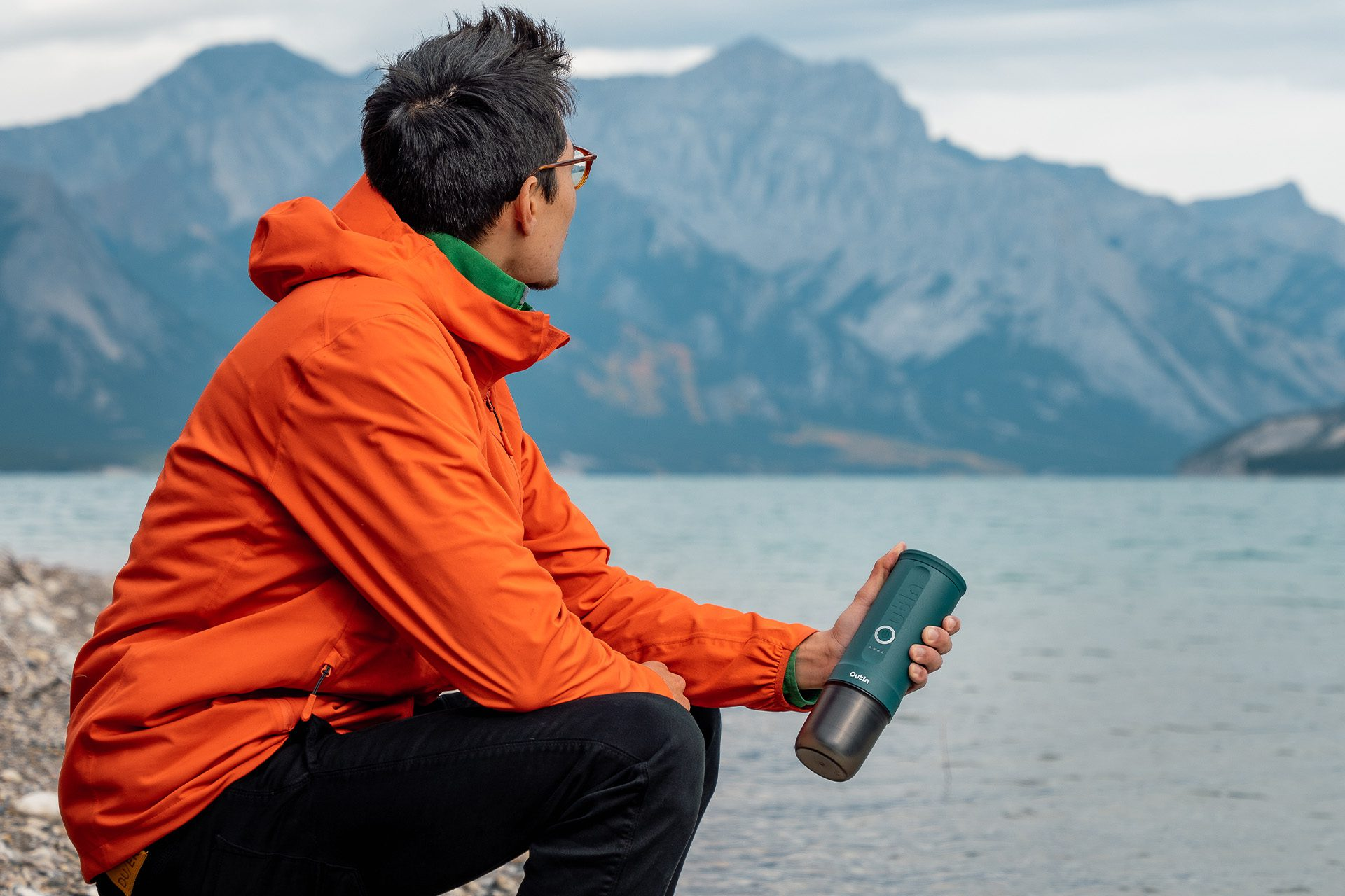 Mężczyzna w pomarańczowej kurtce trzyma zieloną butelkę, patrząc na góry i jezioro. Niebo jest pochmurne.