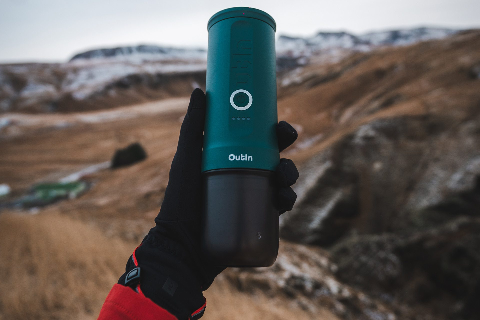 Ręka trzyma tealowo-czarne urządzenie Outin. Tło przedstawia brązowy i ośnieżony krajobraz pod zachmurzonym niebem.