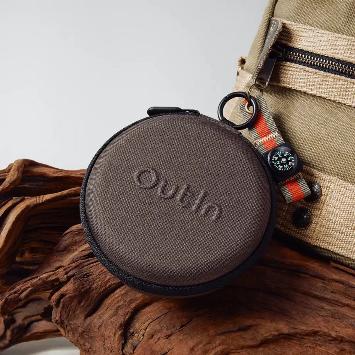 A brown, round case with 'Outln' embossed on it sits on wood. A bag and a compass hang on the right.