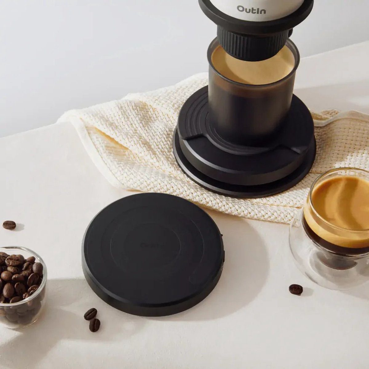 A coffee maker with coffee beans, a cup of coffee, and a black lid on a white surface. A towel is under the coffee maker.