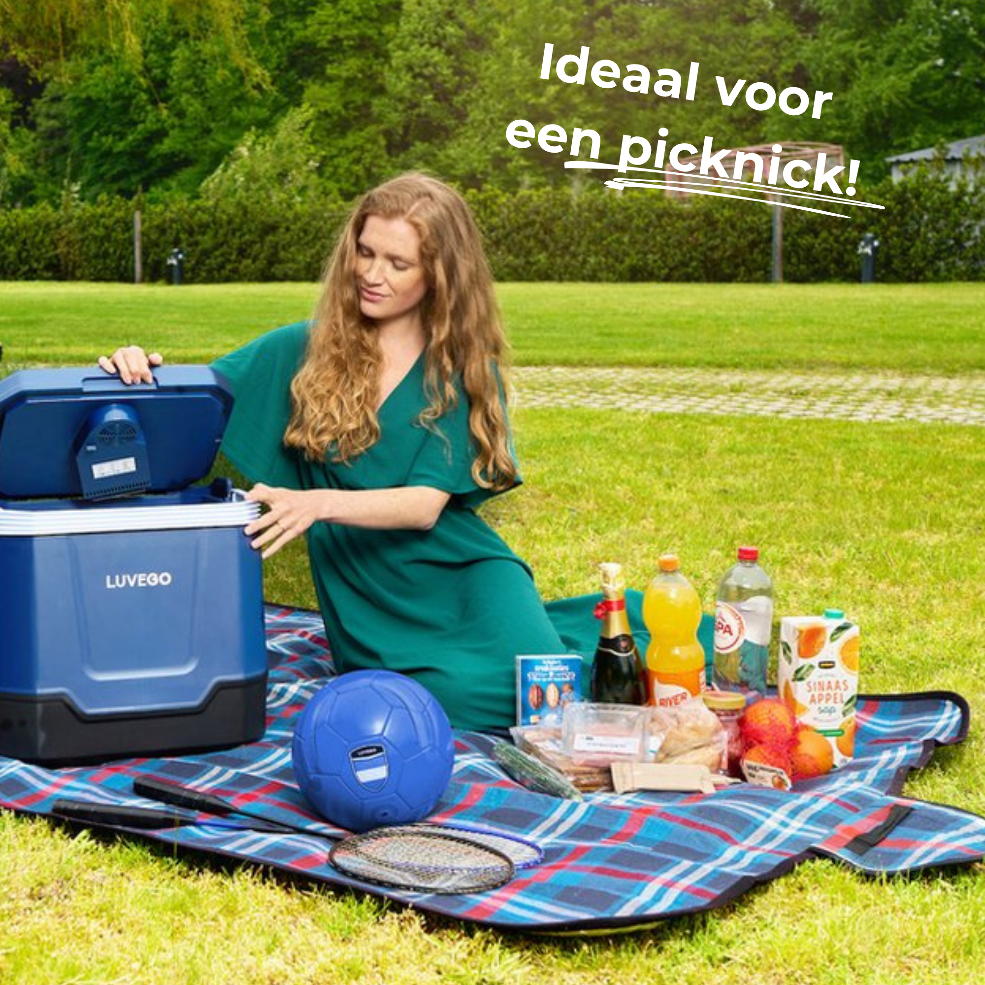 Frau beim Picknick mit Kühler, Essen und Getränken. Blauer Kühler, Fußball und Badmintonschläger sind sichtbar.