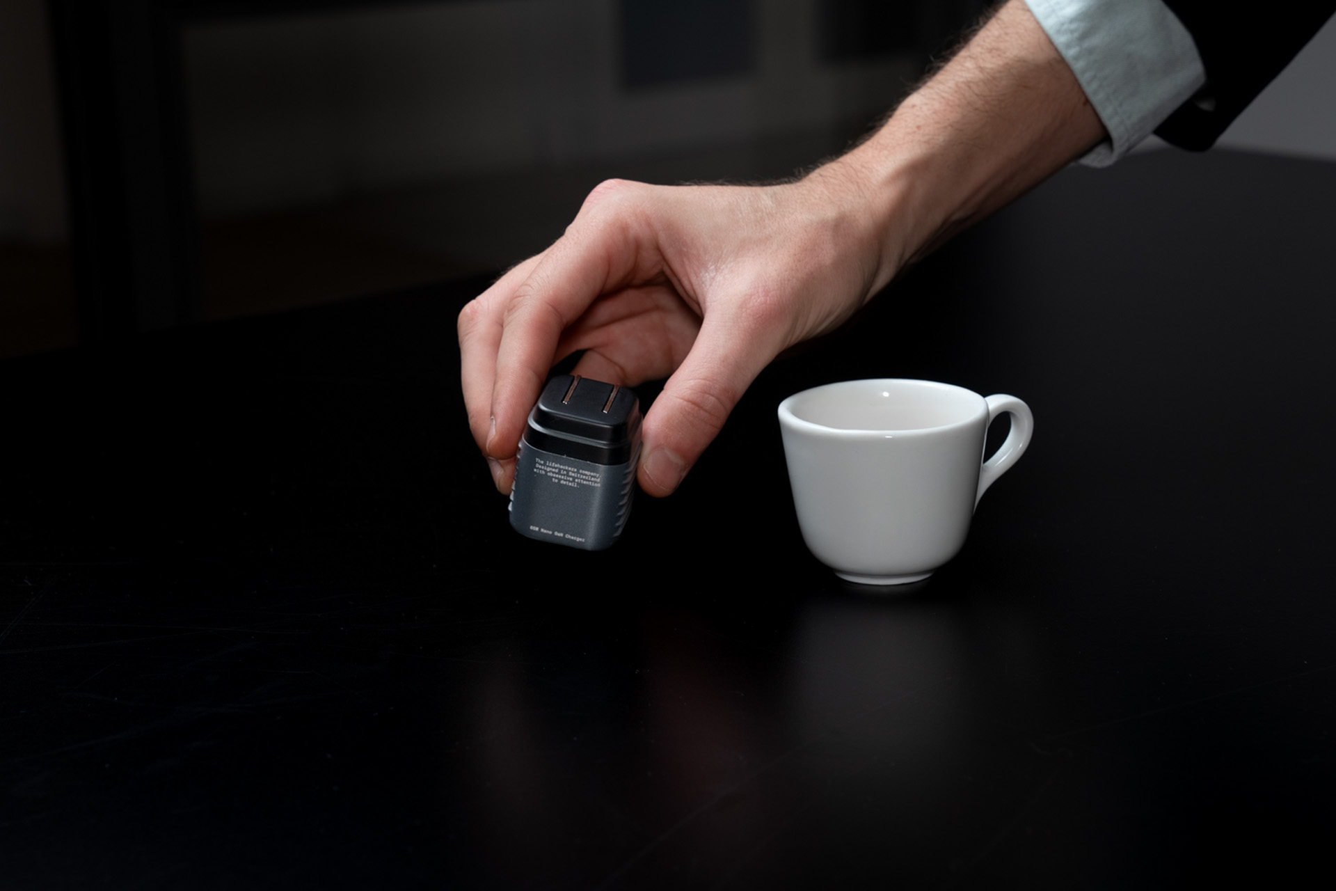 A hand holds a gray device, with a white coffee cup beside it on a black table. A white shirt sleeve is visible.