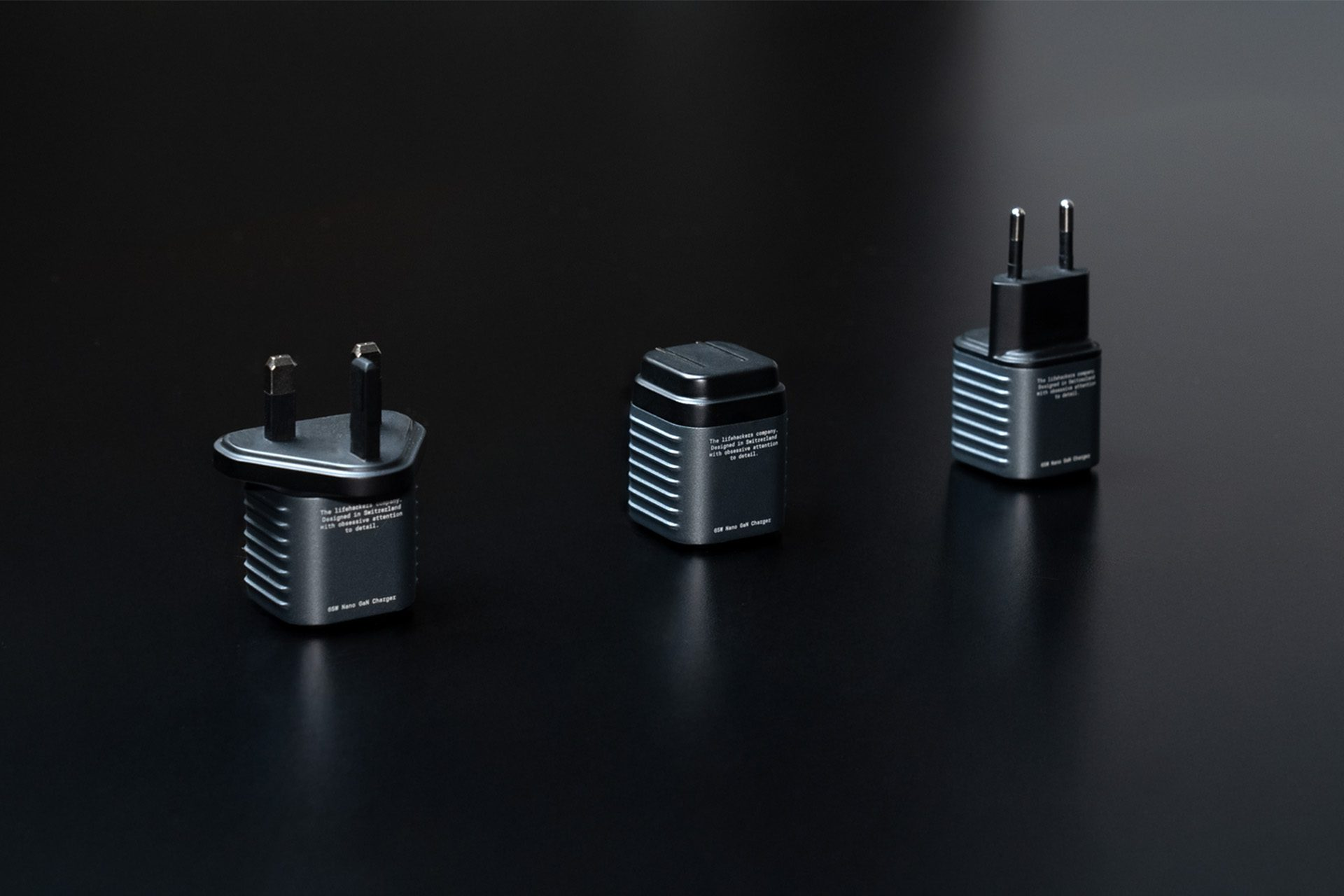 Three dark chargers on a black surface. Each has a different plug type, and text is printed on each charger. The background is solid black.