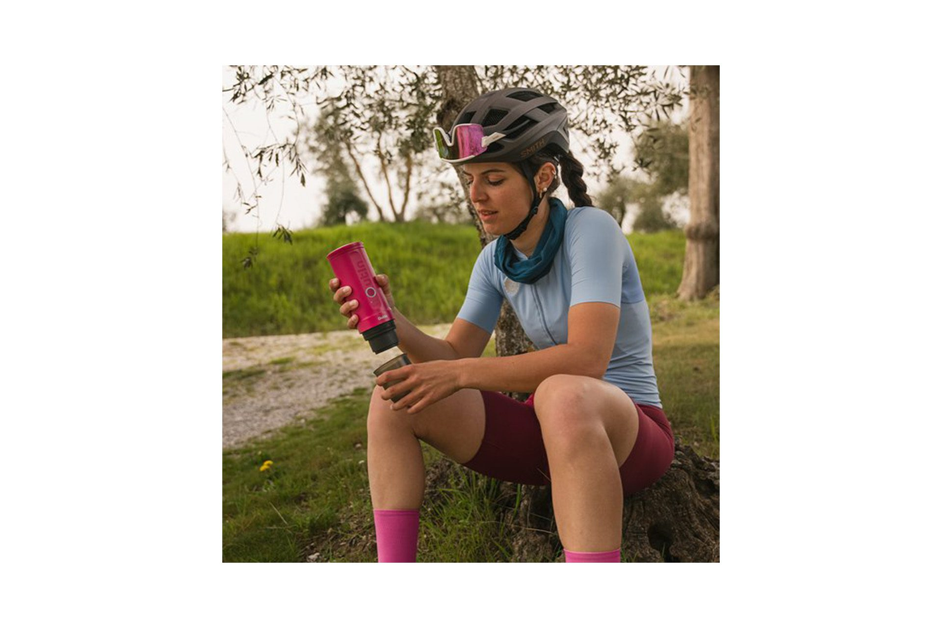 Kolarka siedzi obok drzewa, w kasku i trzyma różową butelkę. Ma na sobie strój kolarski, spodenki i skarpetki.
