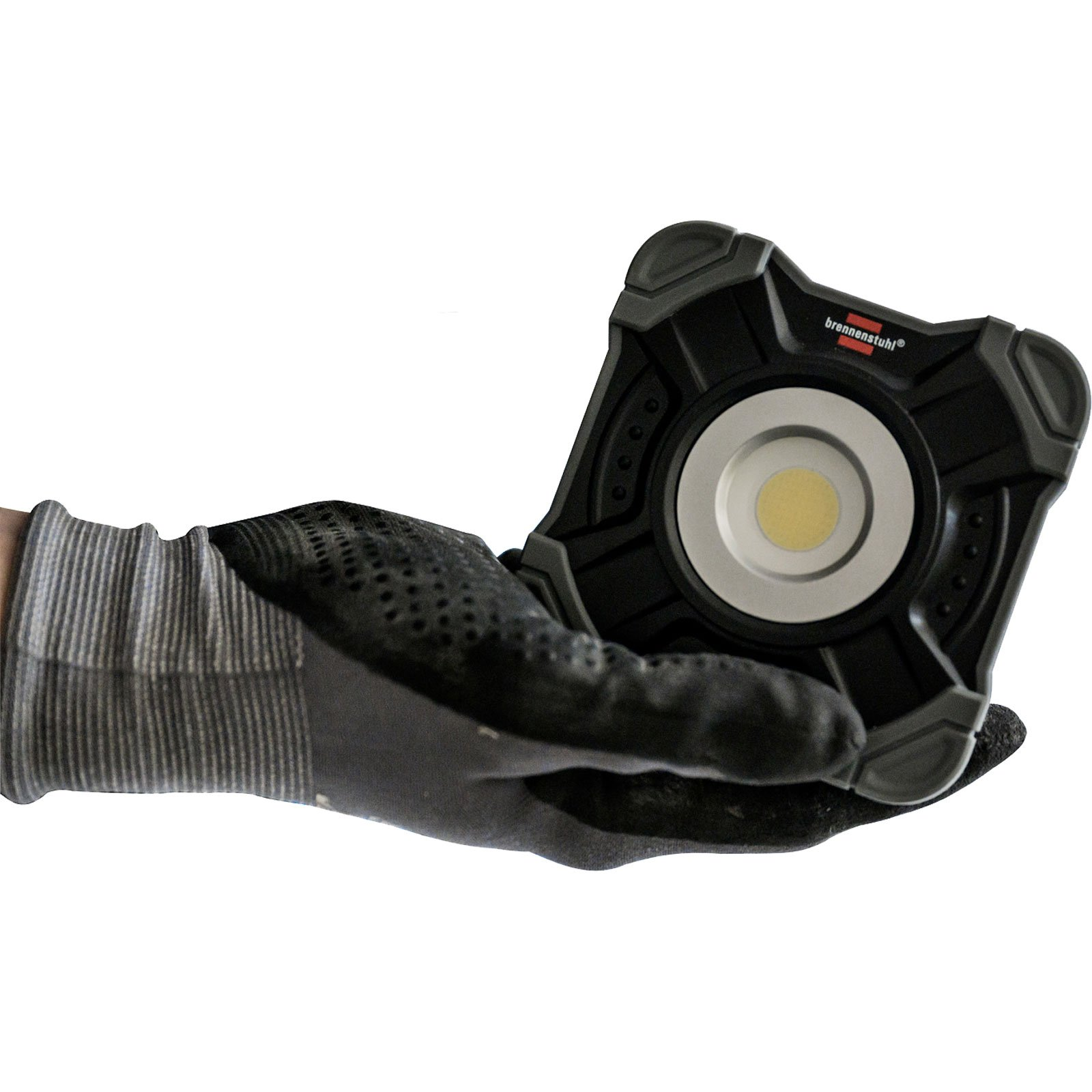 Mano con guanto nero che tiene una lampada da lavoro nera e grigia. La lampada ha un centro luminoso e il marchio 'brennenstuhl'.