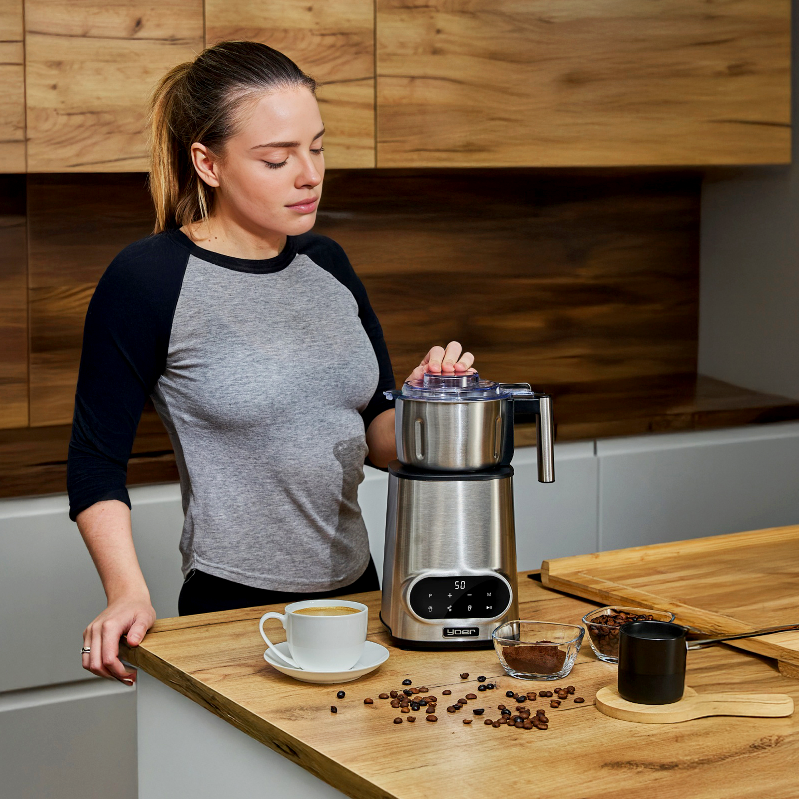 Kobieta obsługuje ekspres do kawy. Filiżanka kawy i ziarna na blacie. W tle szafki kuchenne.